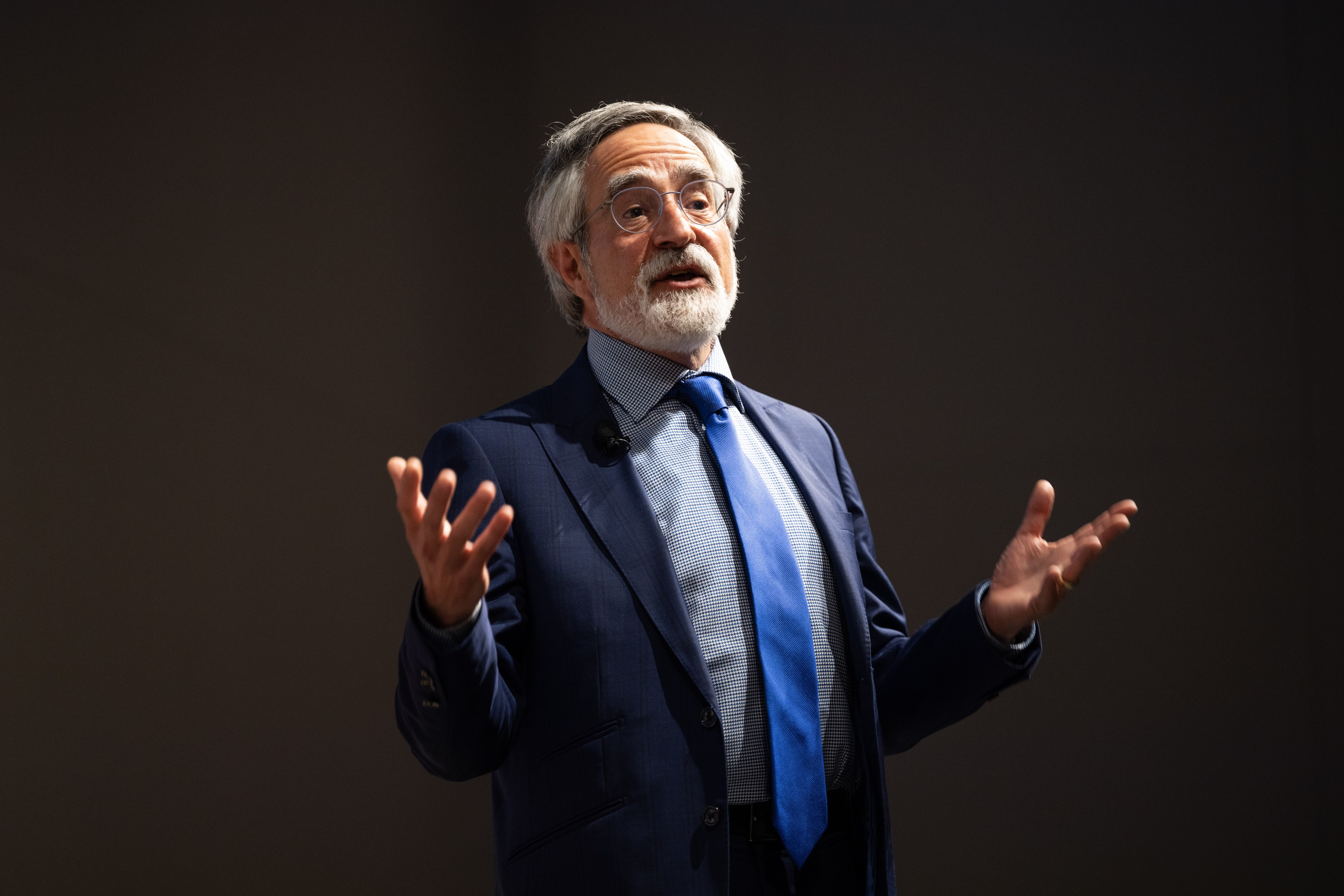 A man with a short gray beard and glasses, wearing a blue suit and tie, is speaking passionately with his hands raised against a dark background.