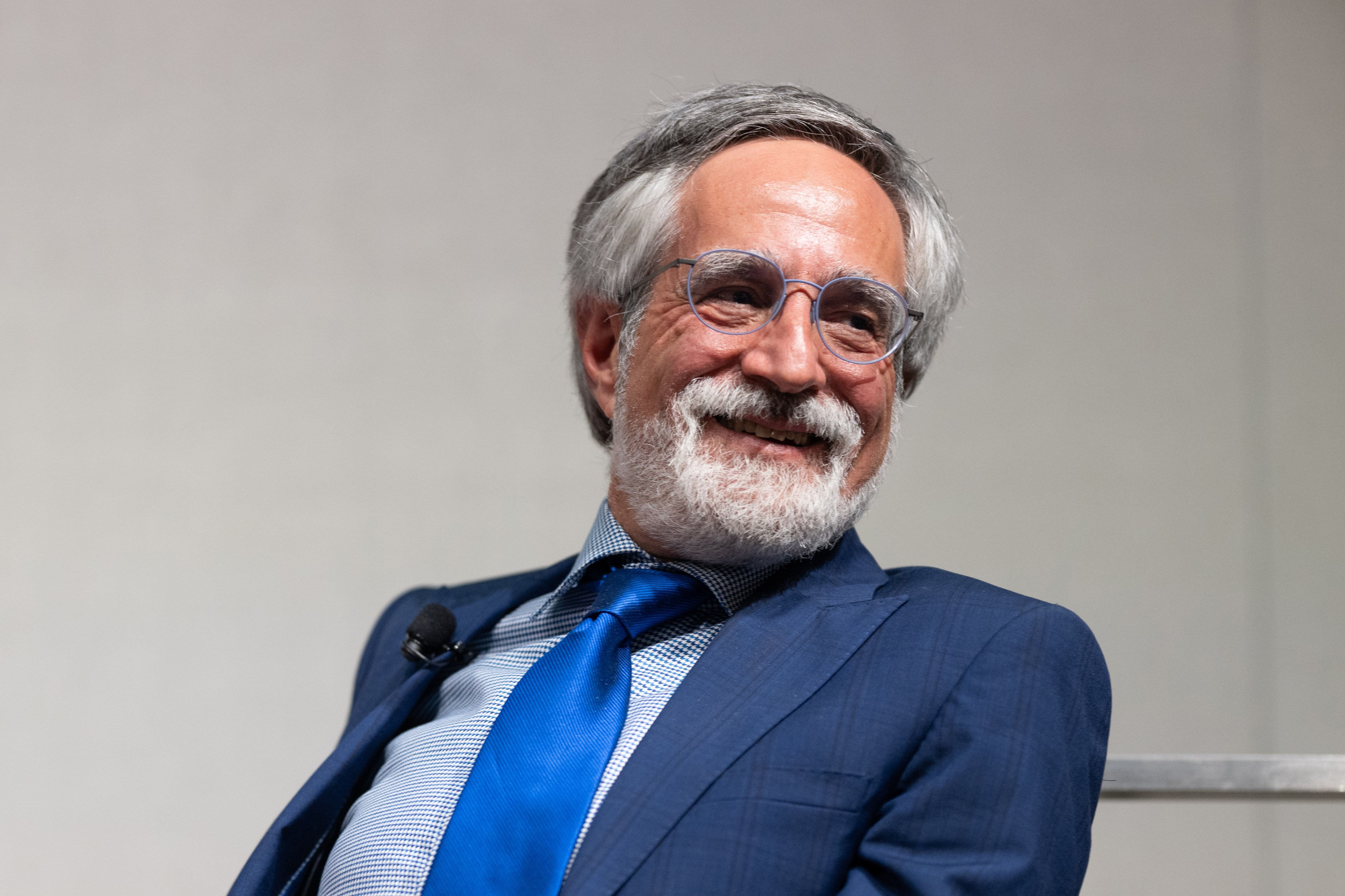 A smiling man with gray hair and beard, wearing glasses, a blue suit, and tie, sits with a microphone clipped to his jacket.