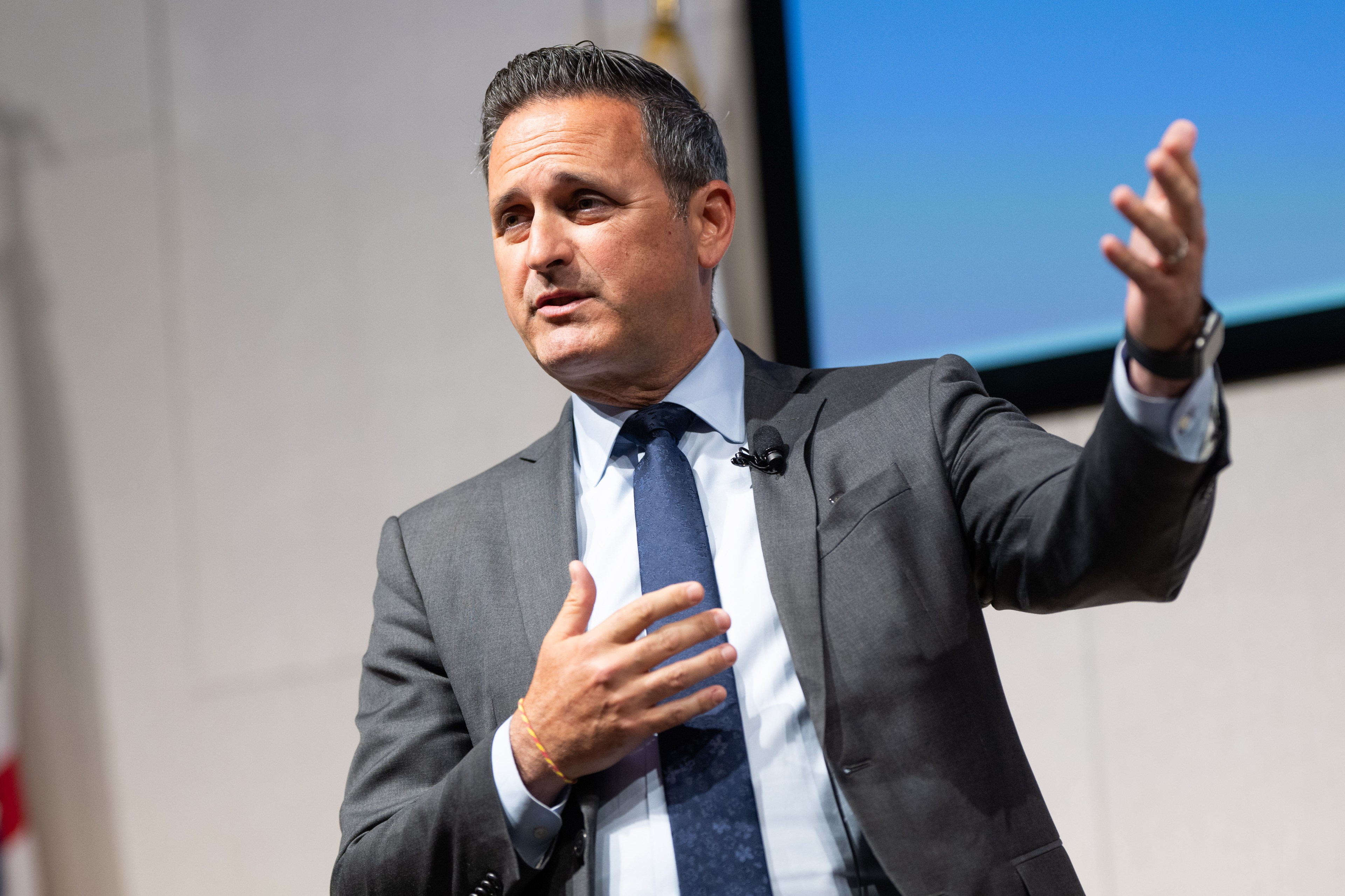 A man in a suit and tie is speaking in front of an audience. He has one hand on his chest and the other gesturing outward. A microphone is clipped to his jacket.