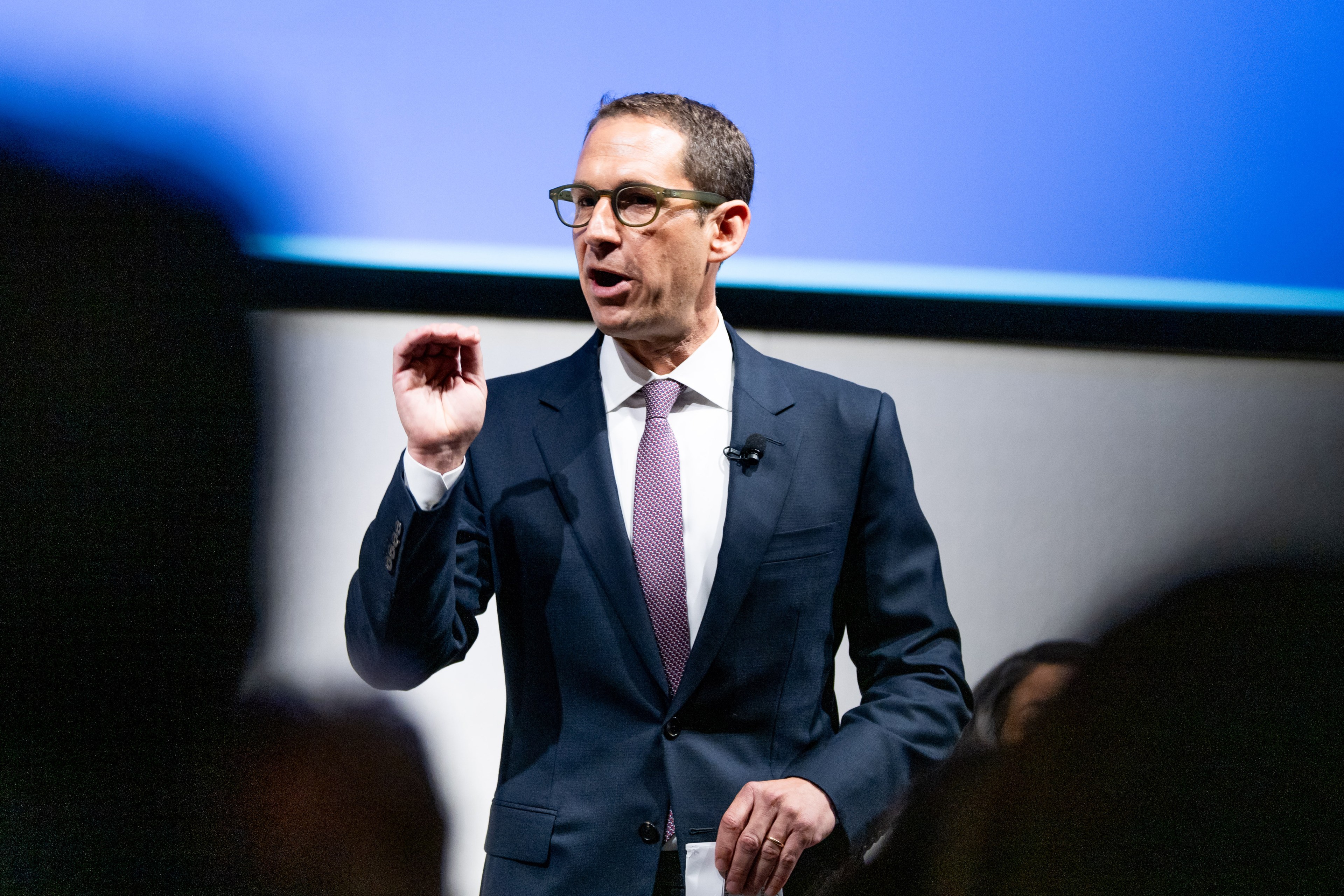 A man in a dark suit and pink tie is speaking on stage, with a blue background and a microphone attached to his jacket.