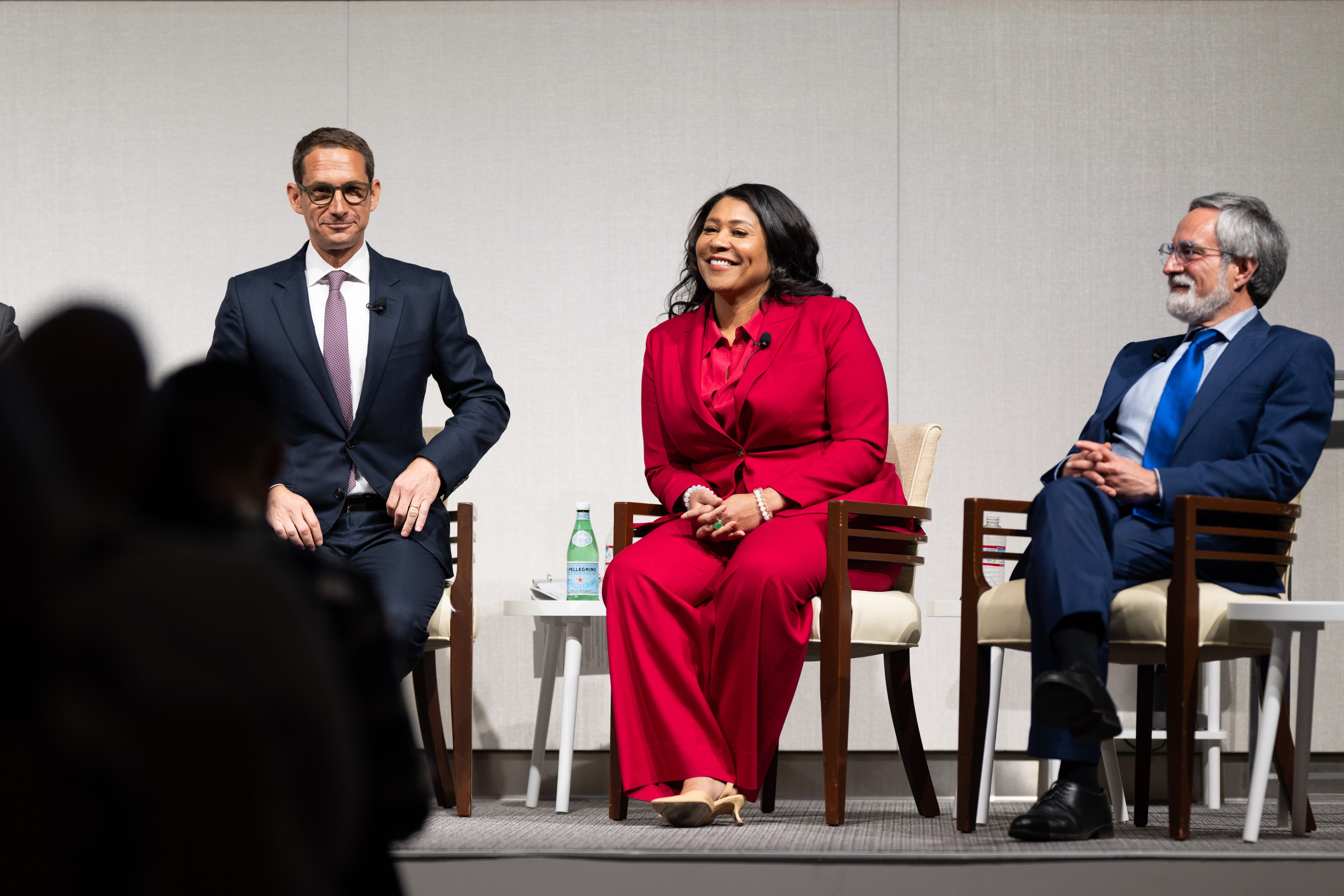 Three people are seated on a stage: a man in a dark suit with glasses, a woman in a red suit smiling, and a man with a beard in a blue suit, all looking attentive.