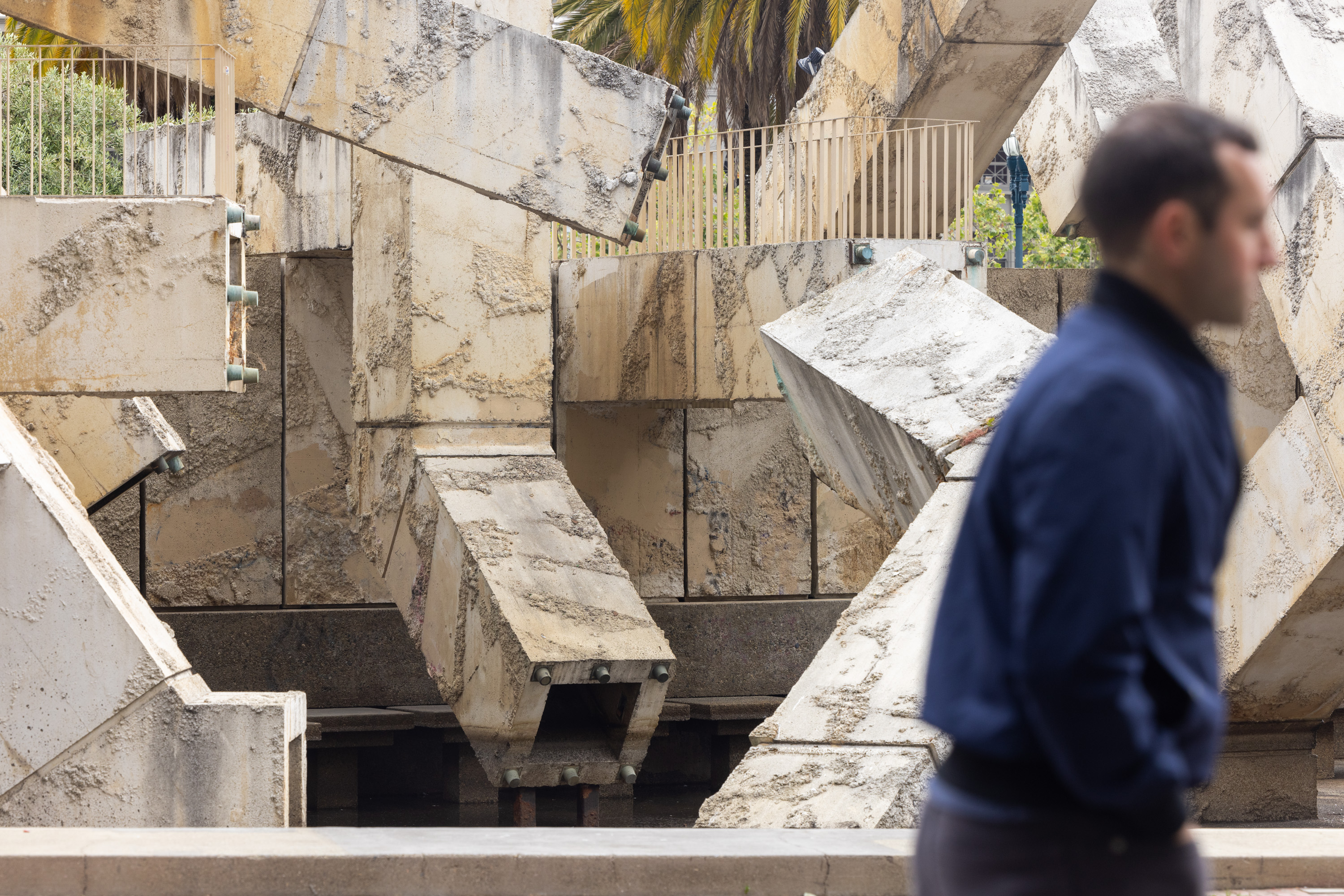 A person in a blue jacket walks past a large, abstract concrete sculpture composed of irregular, weathered geometric shapes, with a palm tree slightly visible in the background.