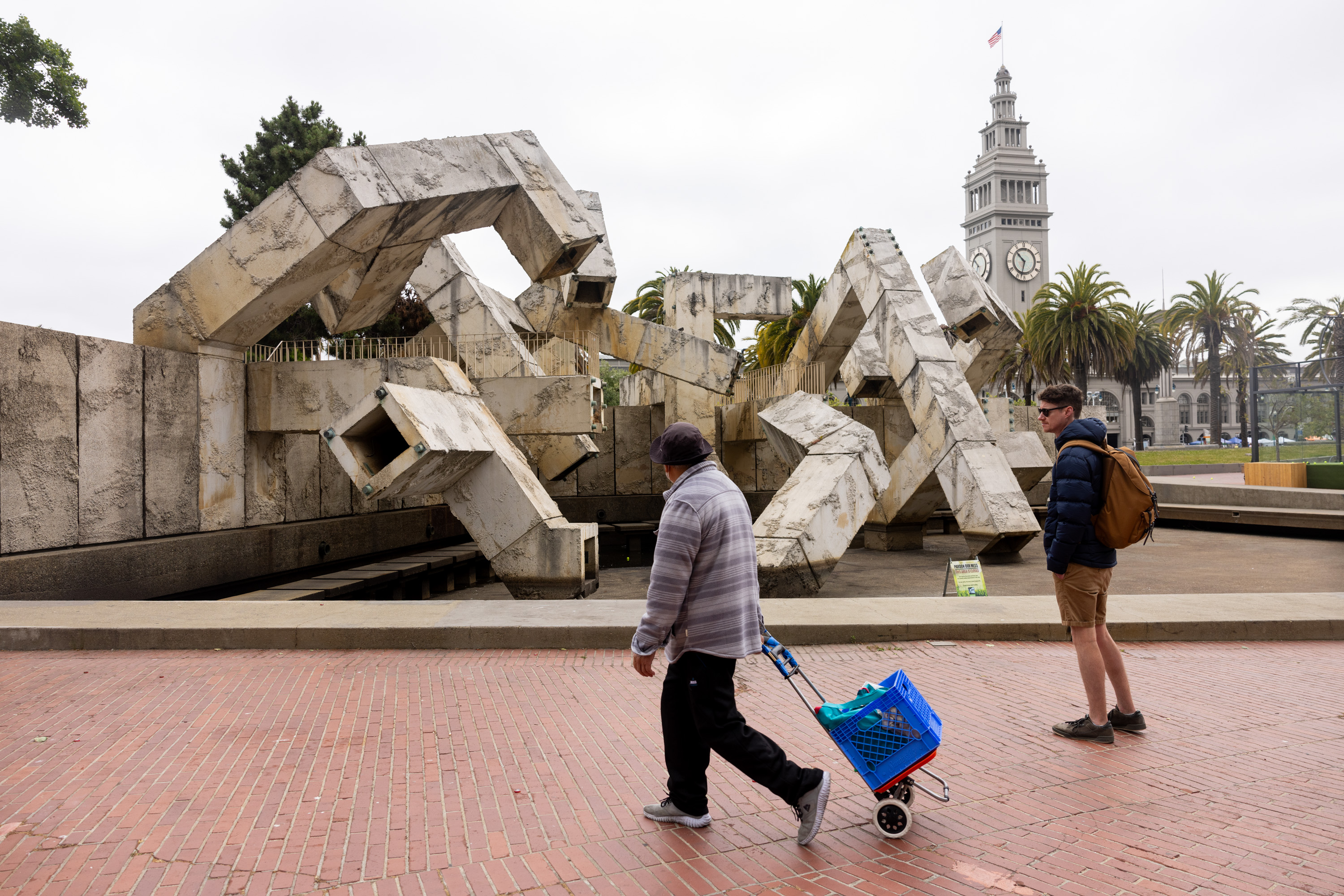 A man with a rolling cart and another person walk by a large, abstract concrete sculpture. In the background, there is a clock tower and palm trees.