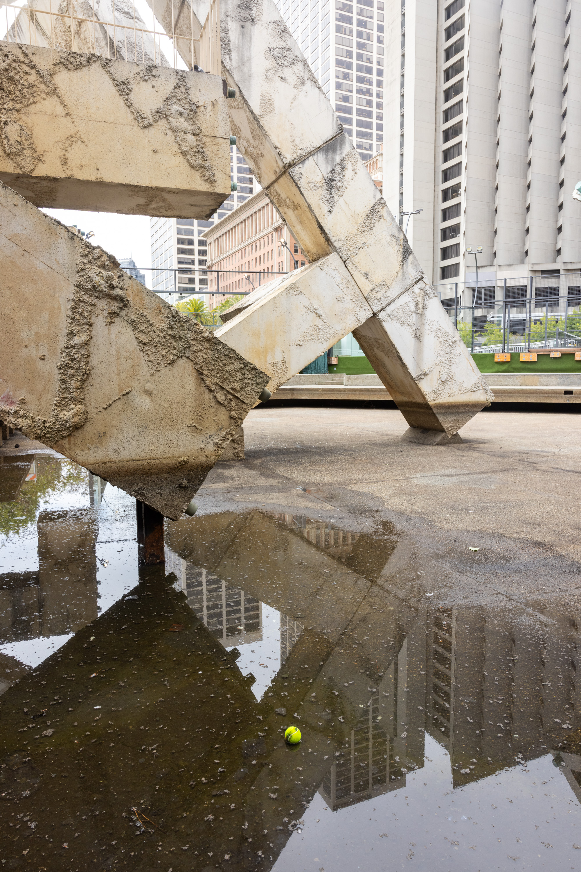 The image shows a large concrete sculpture with rough textures, surrounded by tall buildings. A puddle reflects the sculpture and buildings, with a small green tennis ball in it.