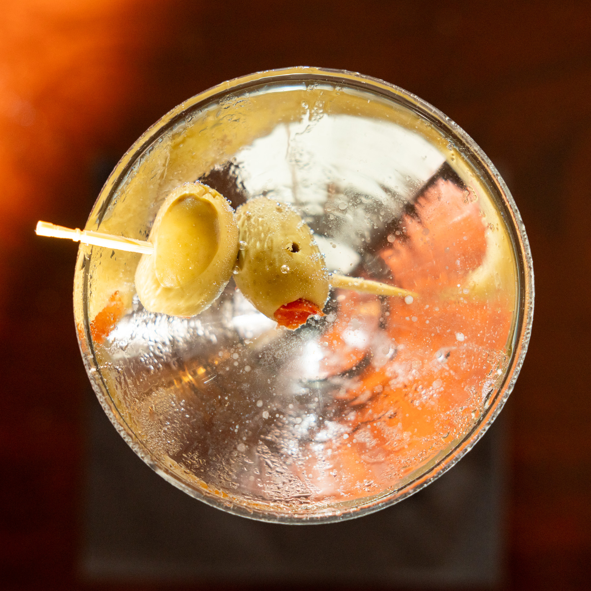 A glass of clear cocktail is garnished with two green olives speared on a toothpick viewed from above. The liquid has condensation on the glass.