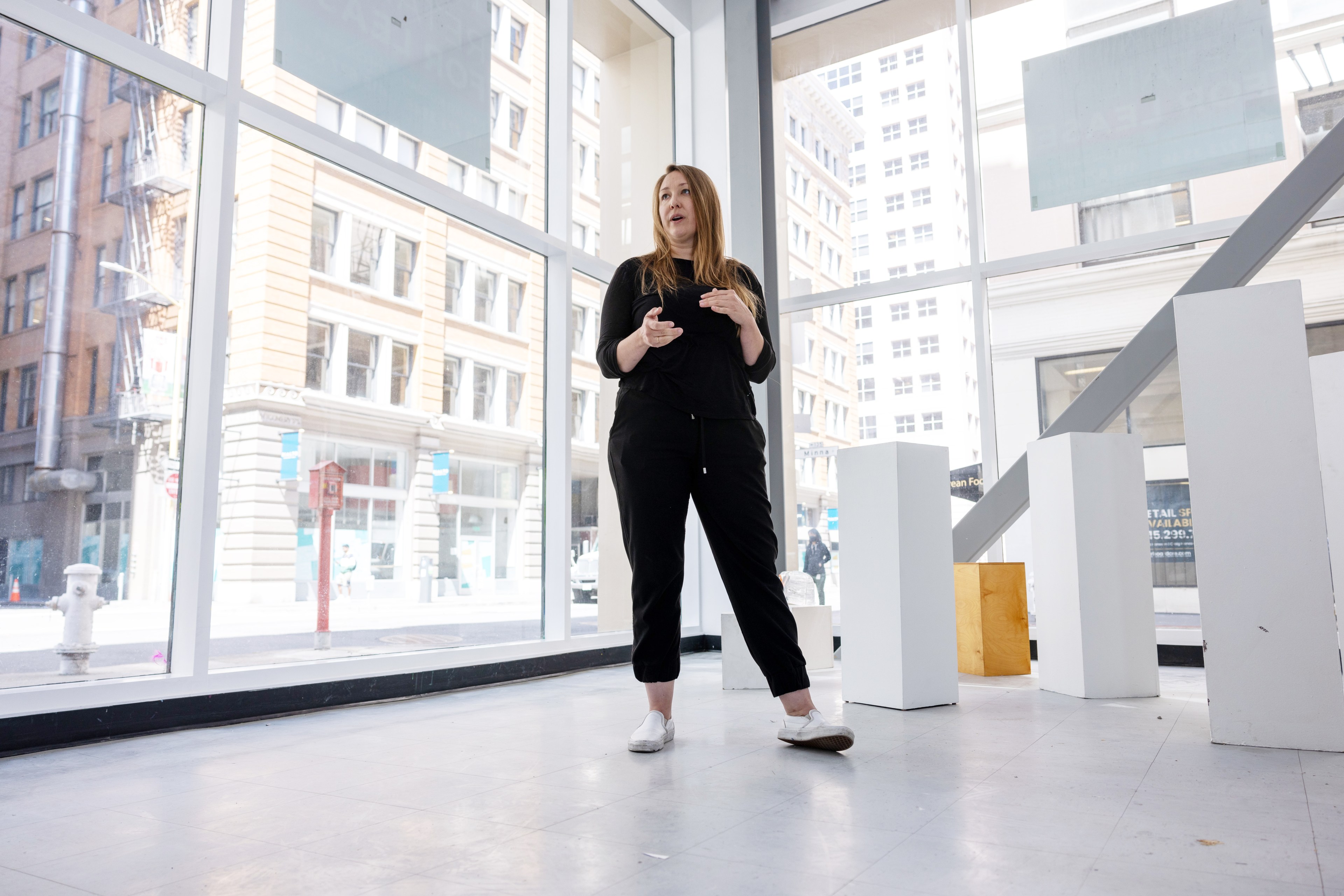 A person with long hair, wearing black clothes and white shoes, is standing and speaking in a large, bright room with large windows and several white pedestals.