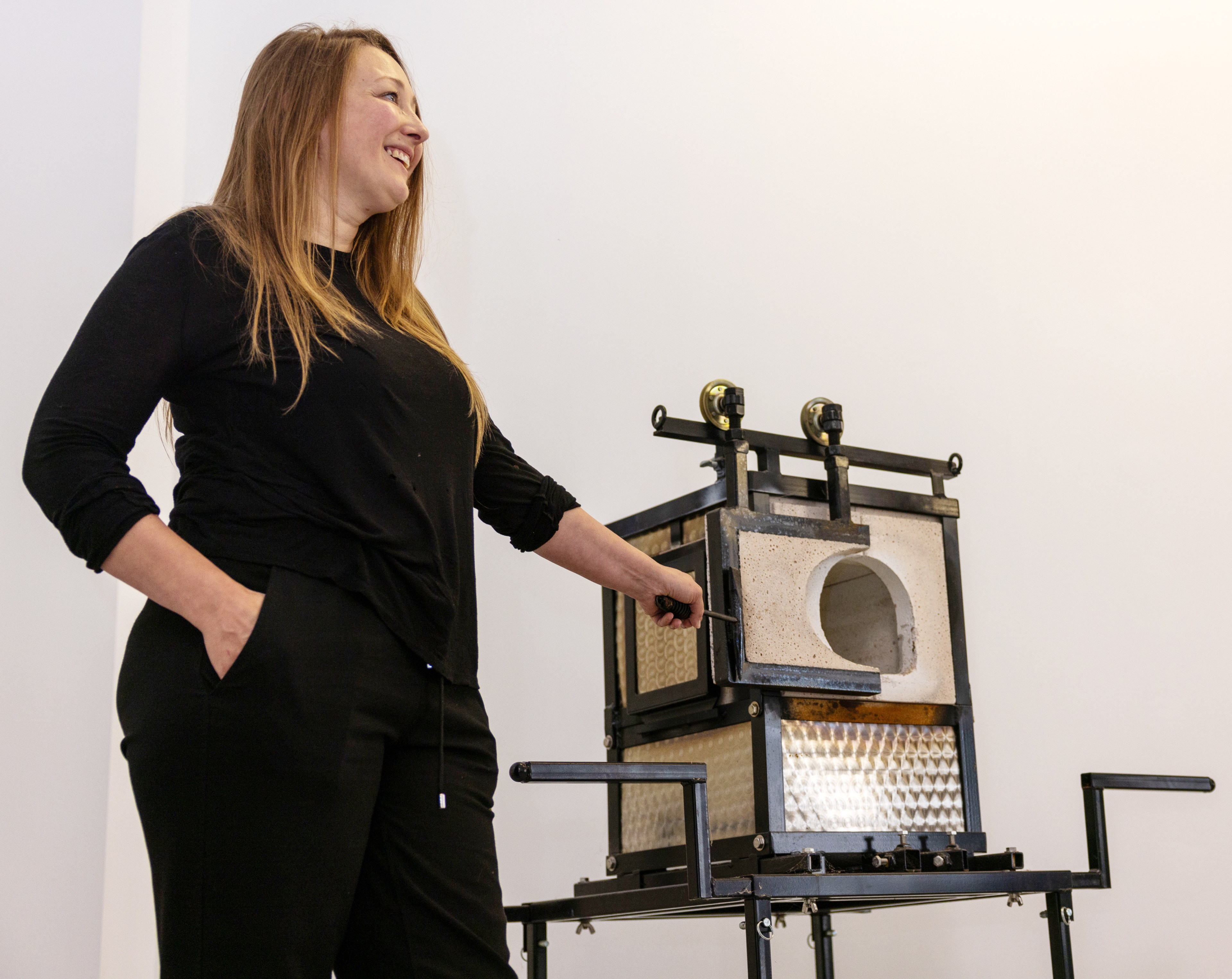 A smiling woman in black attire stands beside a kiln with one hand on her pocket and the other adjusting the kiln's door. The background is plain and white.