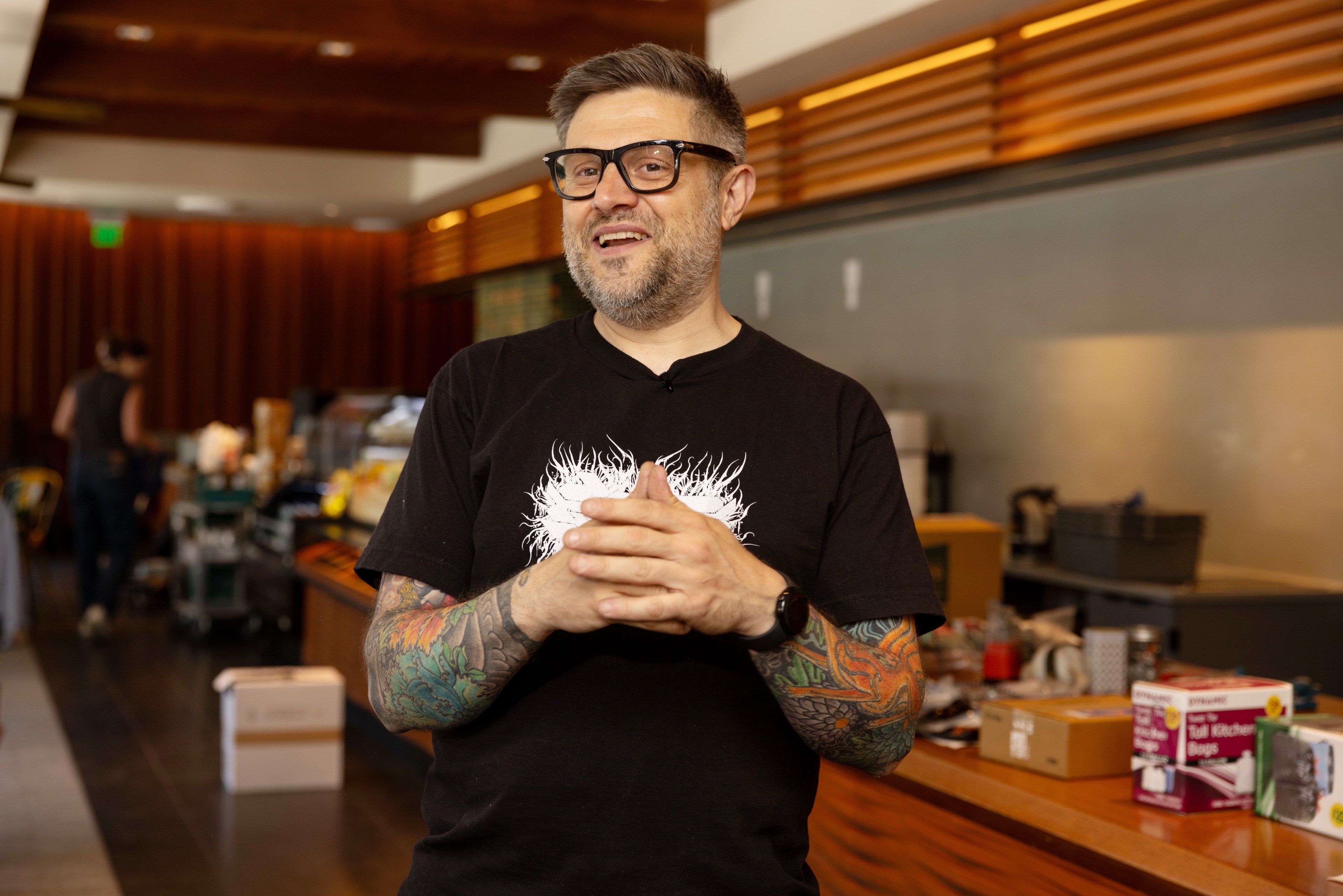 A man with glasses and colorful arm tattoos stands indoors. He wears a black T-shirt with a white design and is speaking, with various items on a counter behind him.