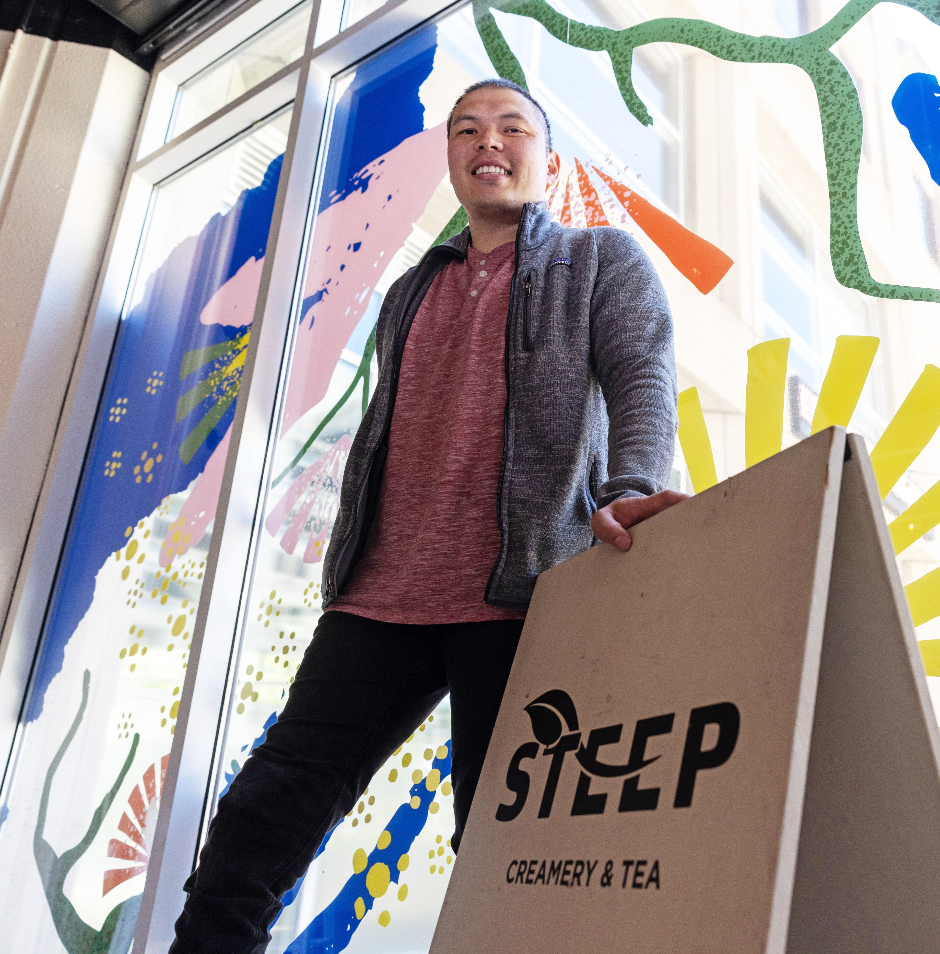 A person is smiling, standing next to a white signboard that reads &quot;STEEP Creamery &amp; Tea.&quot; The colorful window behind features abstract floral designs.