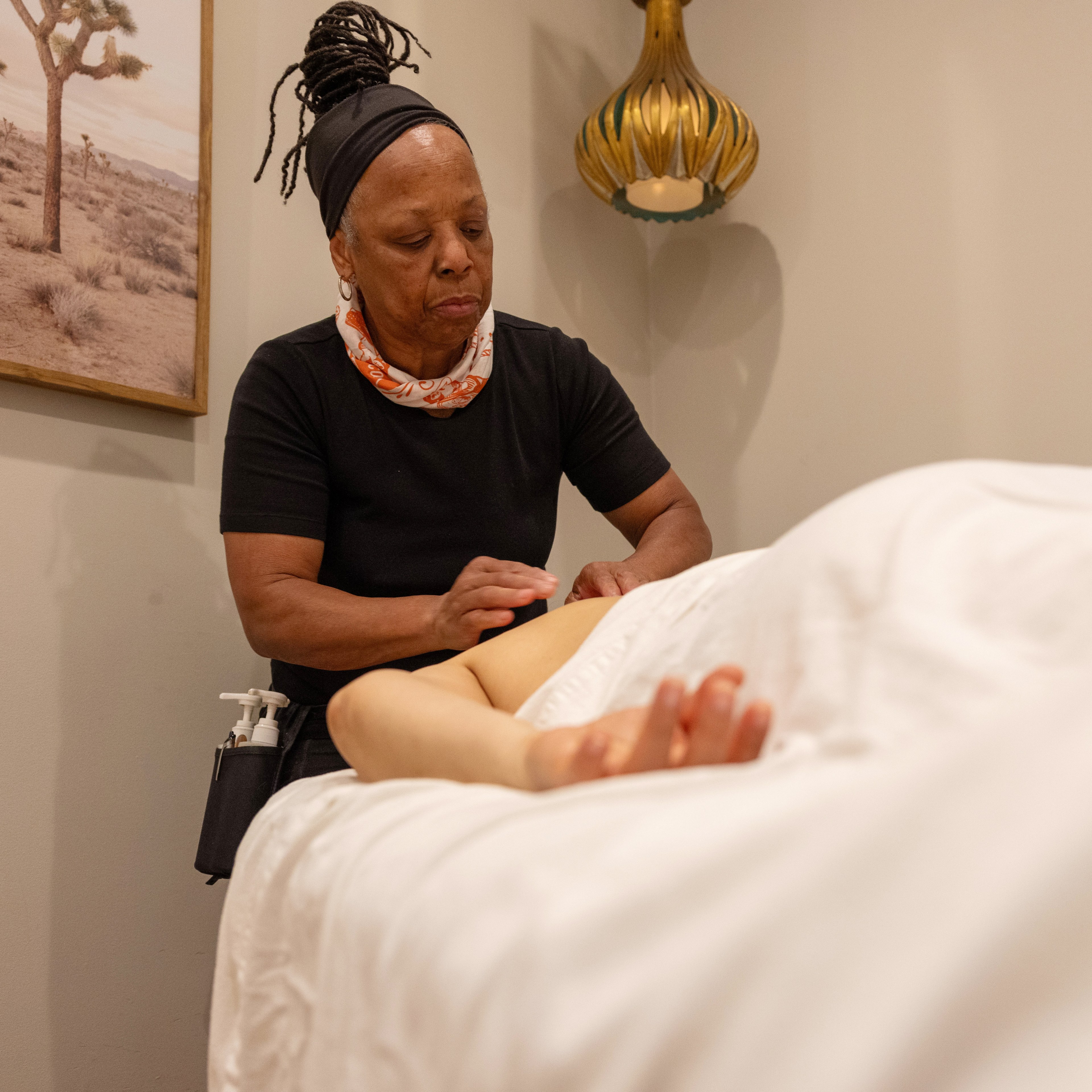 A person is giving a massage to someone lying on a bed, covered with a white sheet. The room has a desert landscape painting and a decorative lamp on the wall.