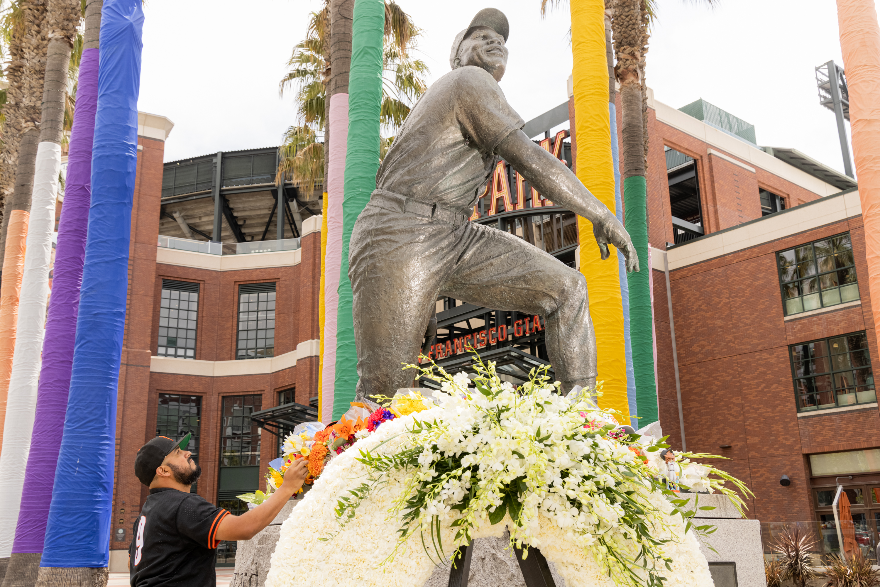 A statue of a baseball player is surrounded by colorful wrapped poles and flowers. A man wearing a baseball jersey is adjusting a floral display at the statue's base.