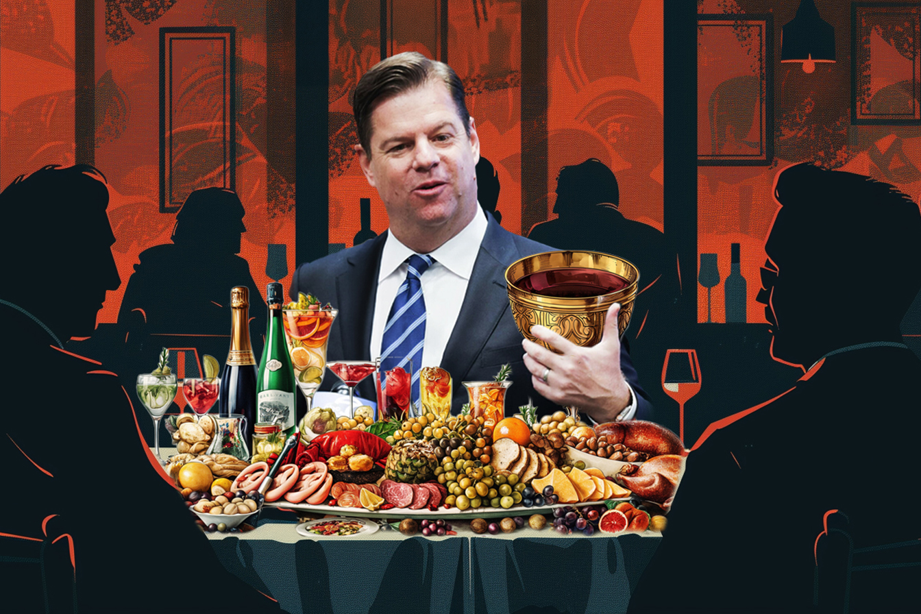 Mark Farrell in a suit holds a golden cup at a lavish table with meats, cheeses, fruits, and drinks, while silhouetted figures sit around in a dimly lit dining setting.