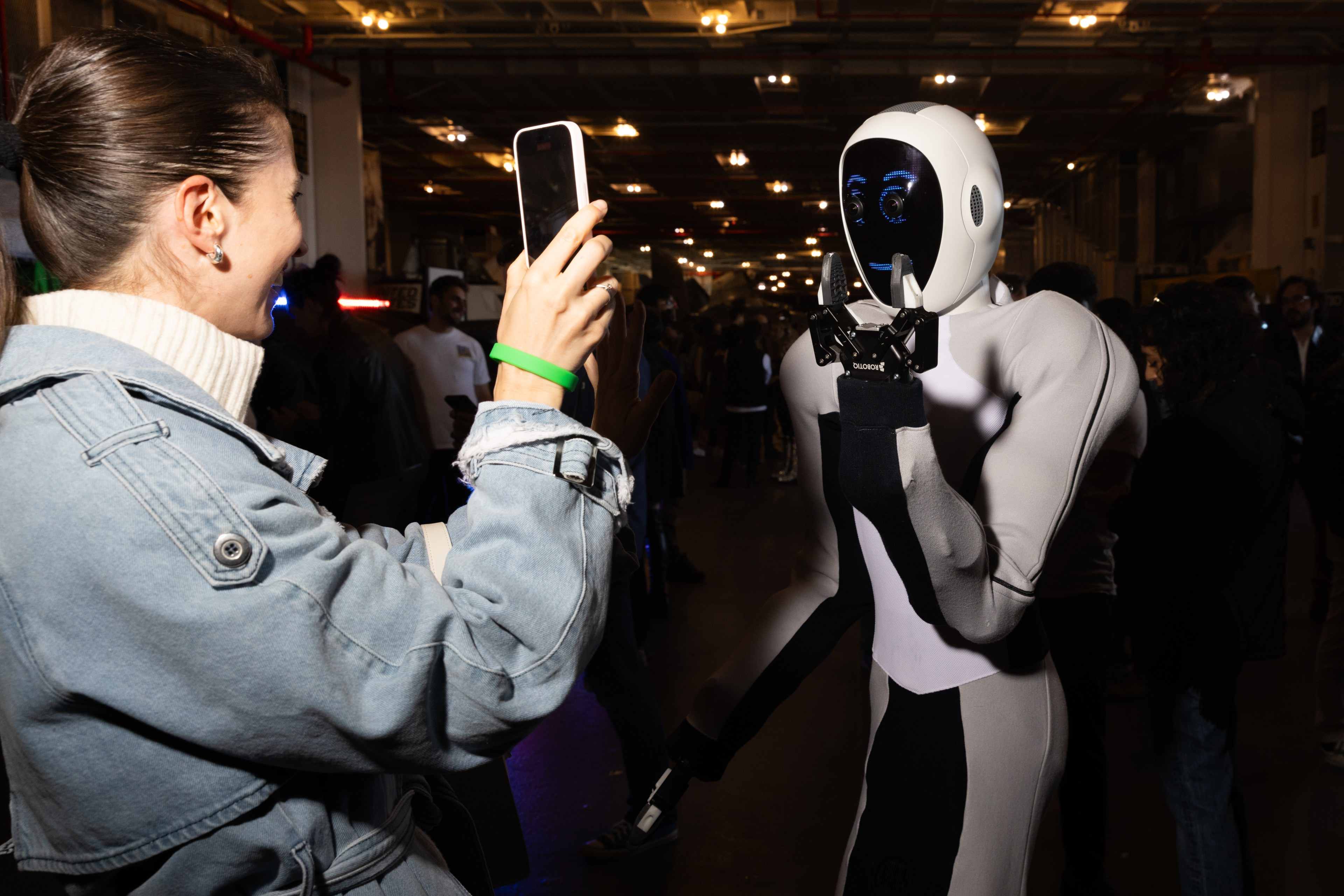 A woman takes a photo of a humanoid robot with her smartphone in a dimly lit indoor setting where several people are present in the background.