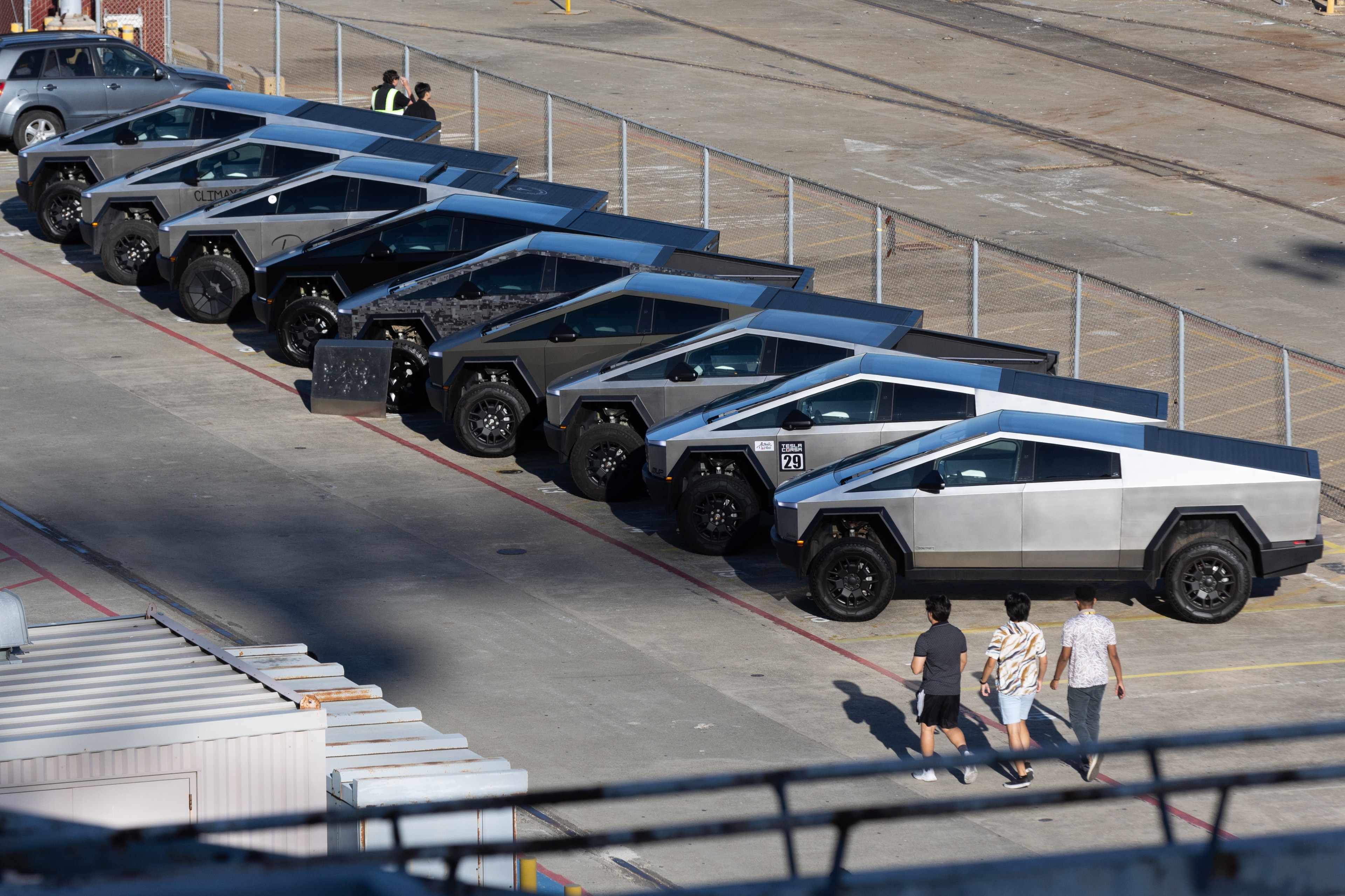 The image shows a row of futuristic, angular truck-like vehicles parked in a lot. Three individuals are walking nearby. The area is fenced and has an industrial feel.