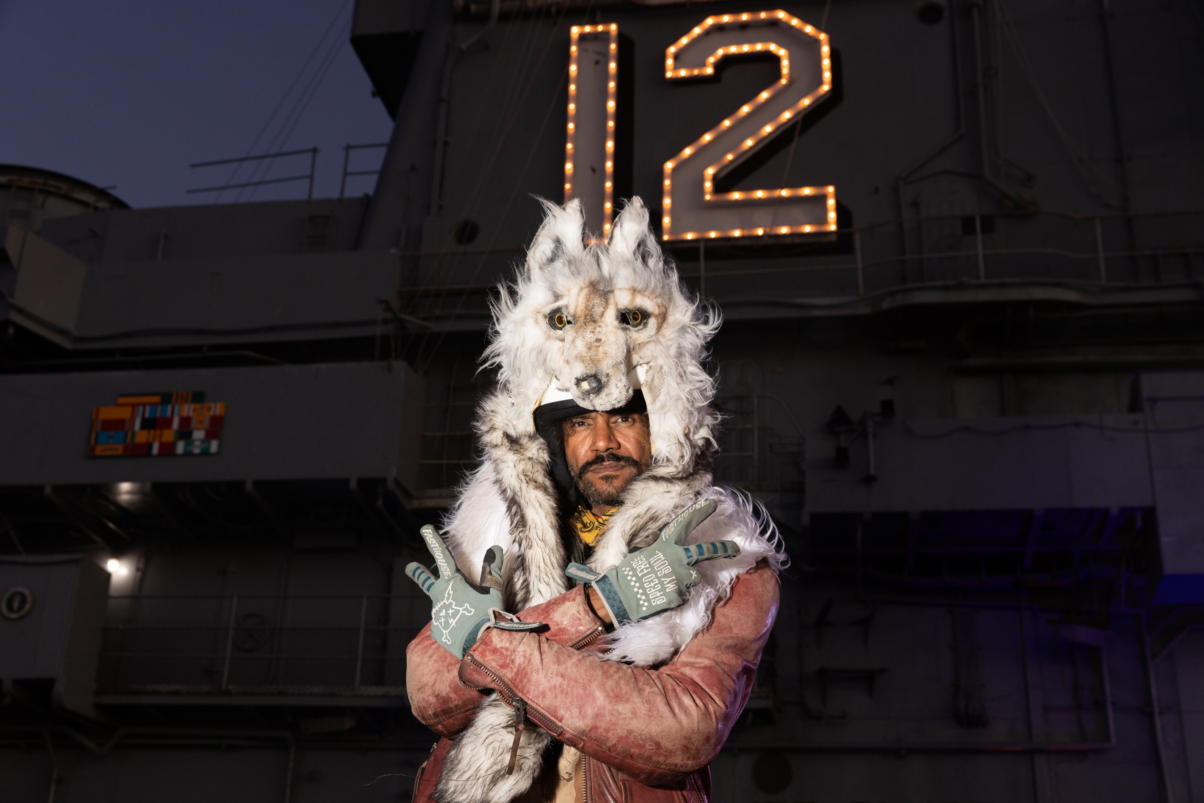 A person wearing a wolf-themed costume stands in front of a structure illuminated by a large number "12." They pose with crossed arms and gloved hands forming gestures.