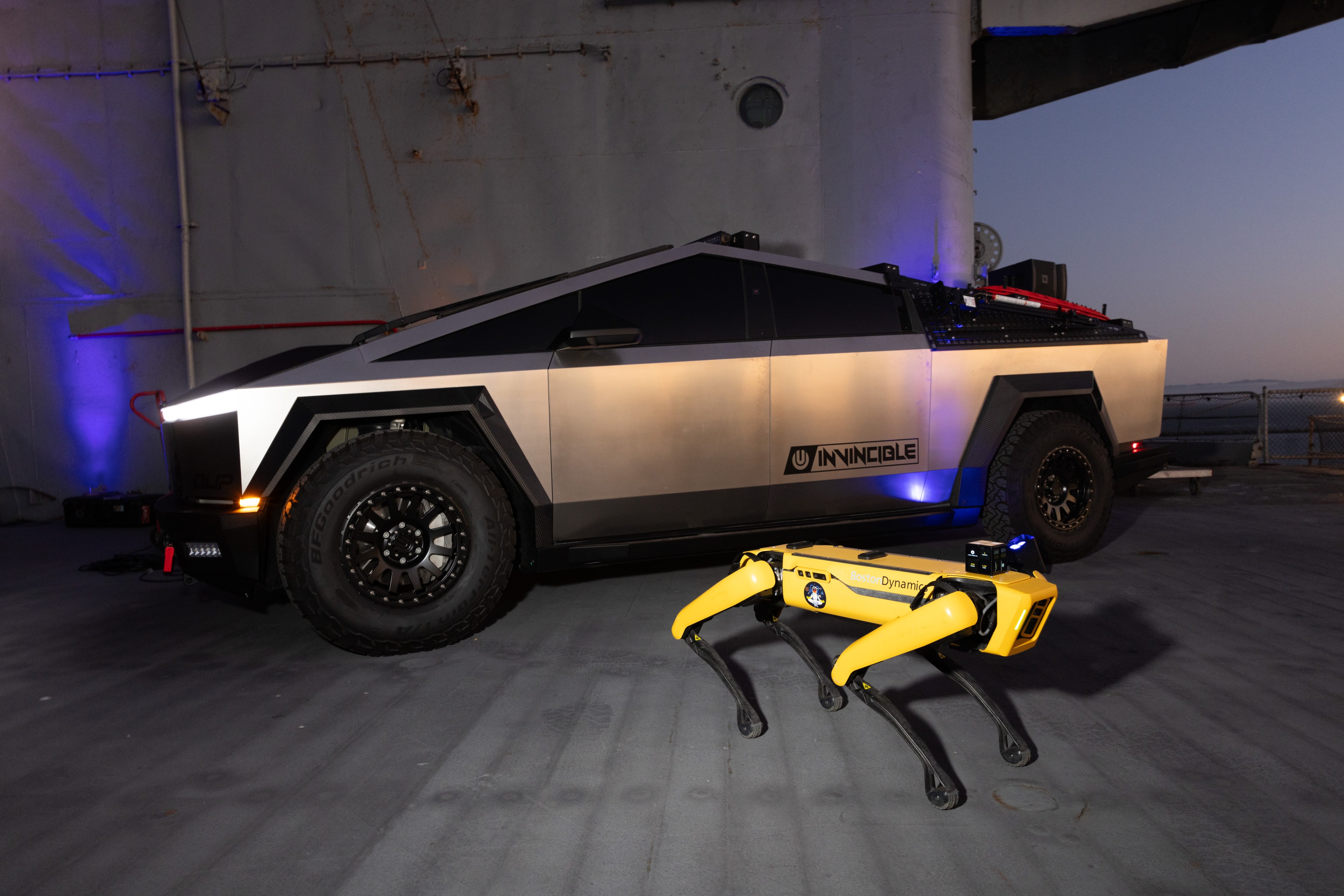 A futuristic silver truck with angular design and large tires is parked indoors next to a yellow, four-legged robotic device on a gray floor.