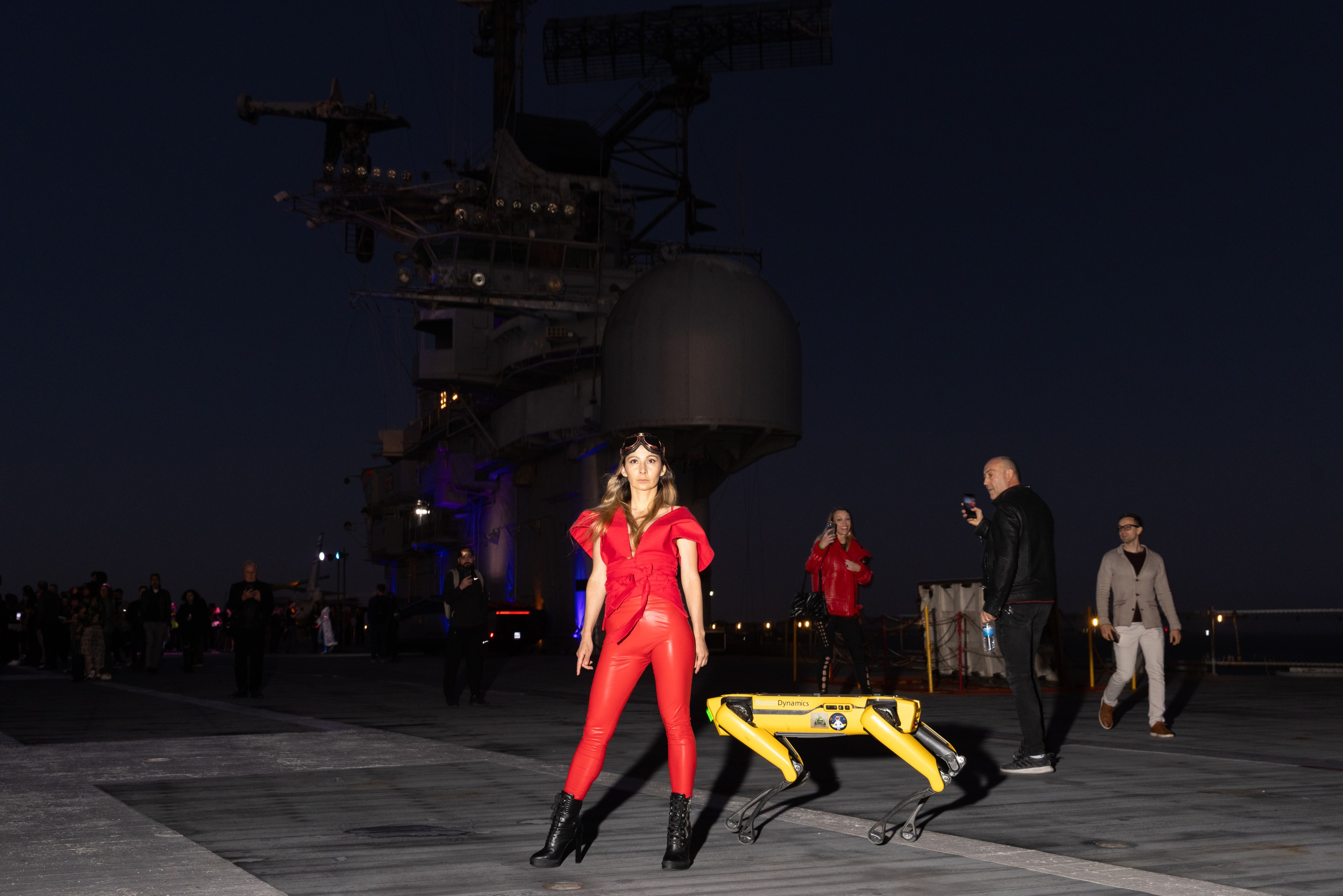 A woman in bright red attire poses confidently next to a yellow robotic dog on an outdoor platform at night; bystanders observe and photograph the scene.
