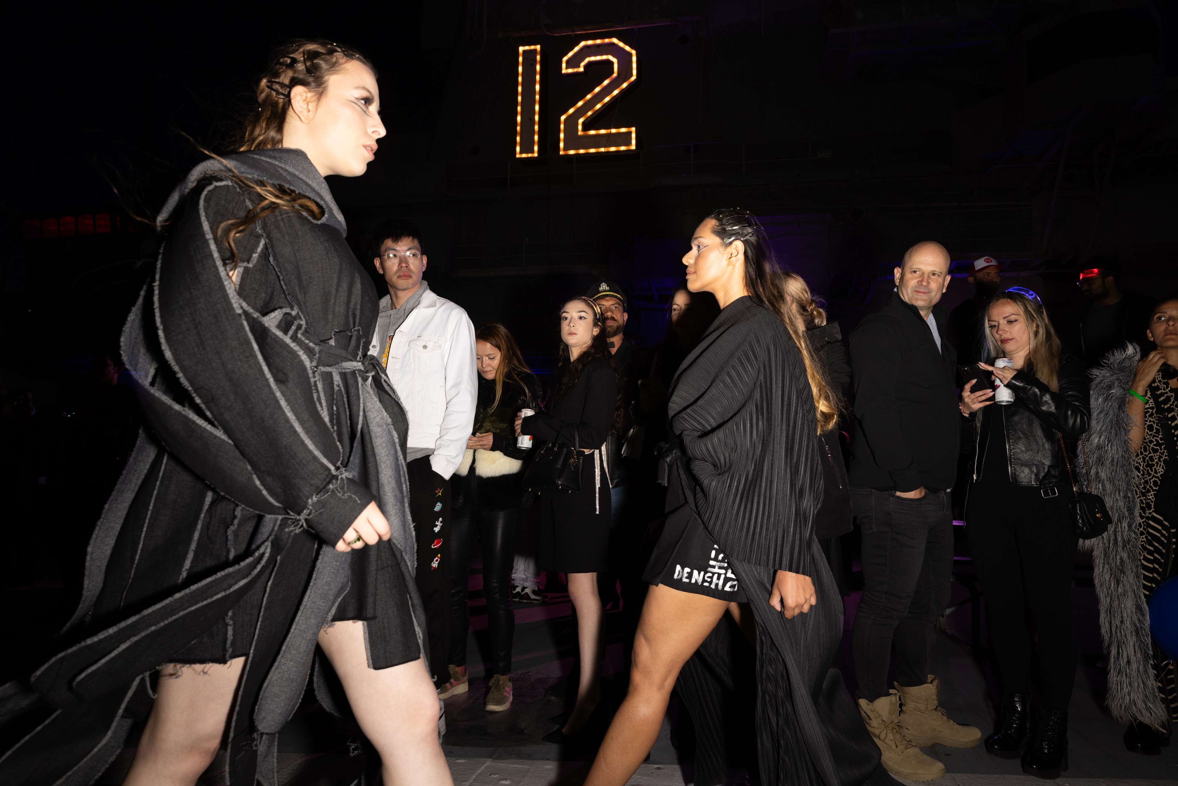 Two models in black outfits walk a runway while an audience watches. The background is dark with a &quot;12&quot; light display above. Some people are engaged and others look at their phones.