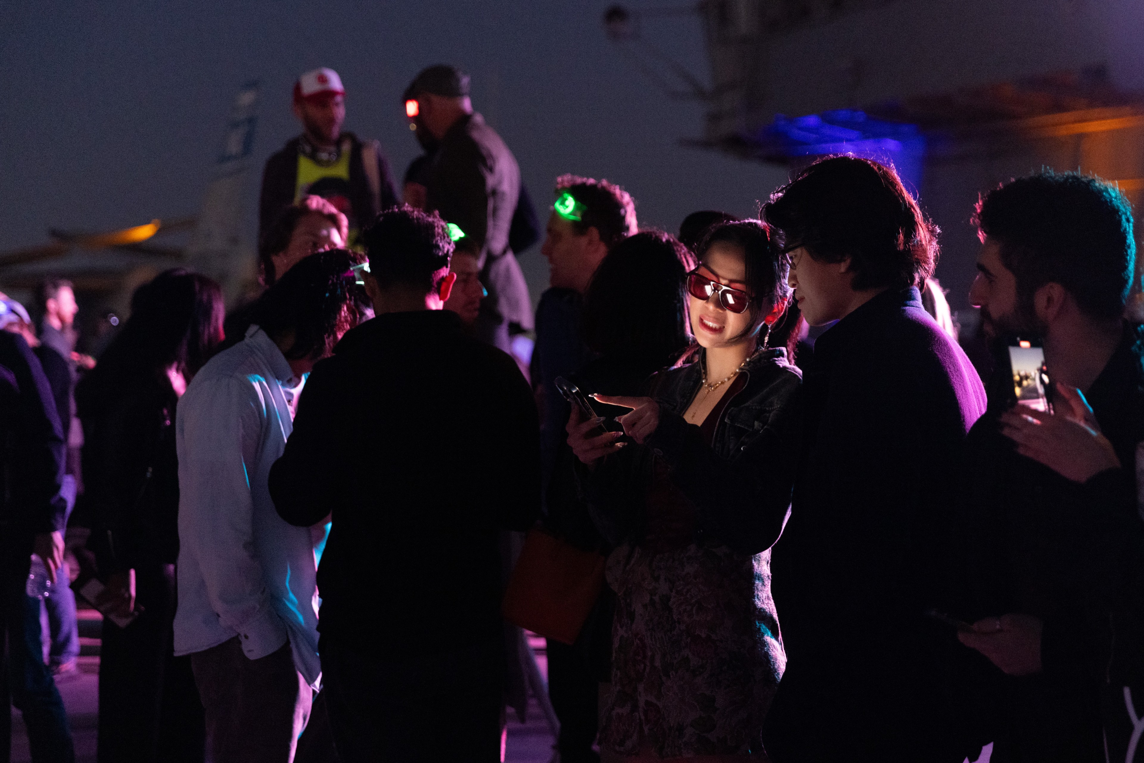 A group of people is gathered at an evening event, with many wearing sunglasses and some illuminated by colorful lights. A couple appears to be sharing something on a phone.