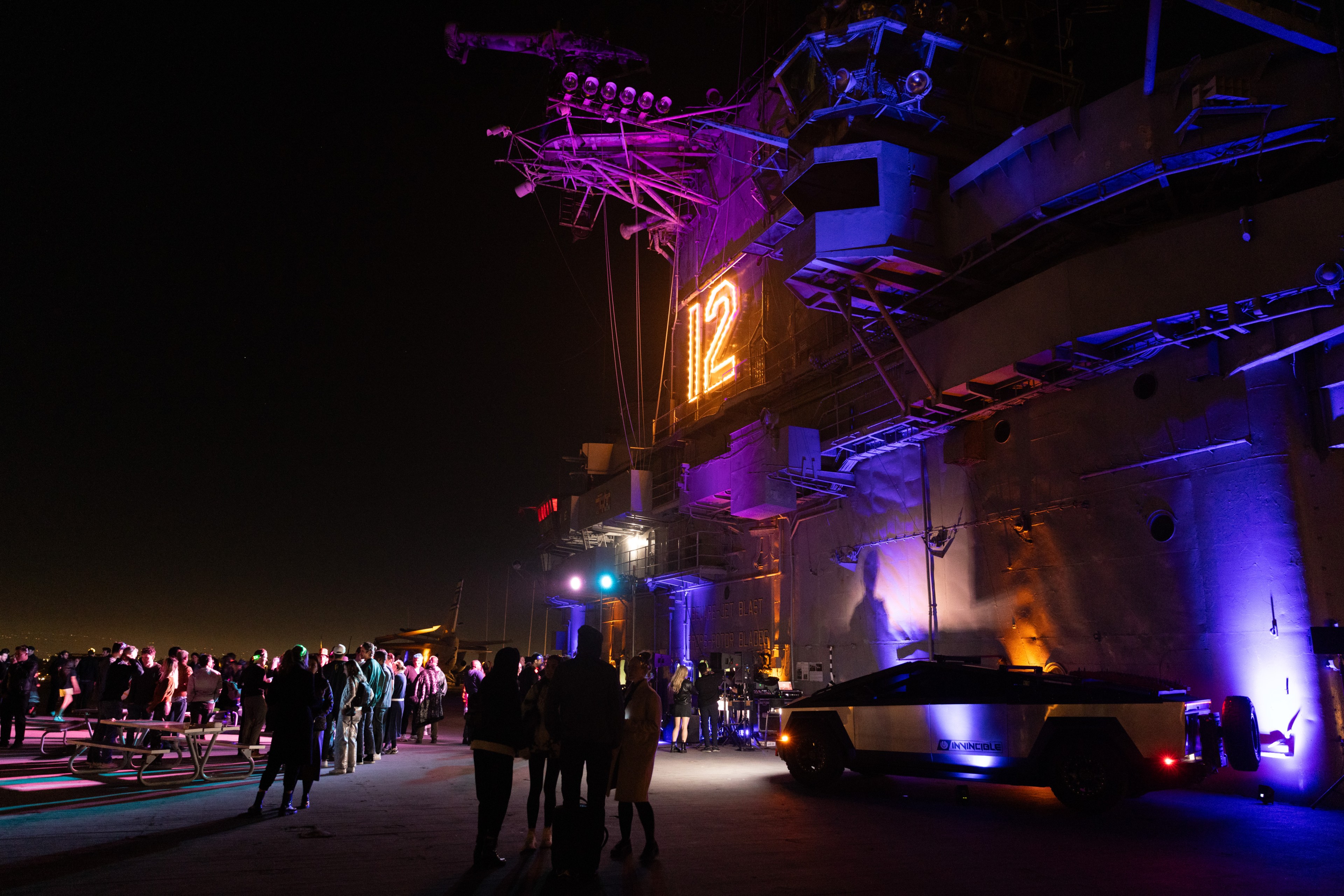 People gather at night on a ship's deck with colorful lights. A lit &quot;12&quot; is displayed on the structure, and a distinctively designed vehicle is illuminated nearby.