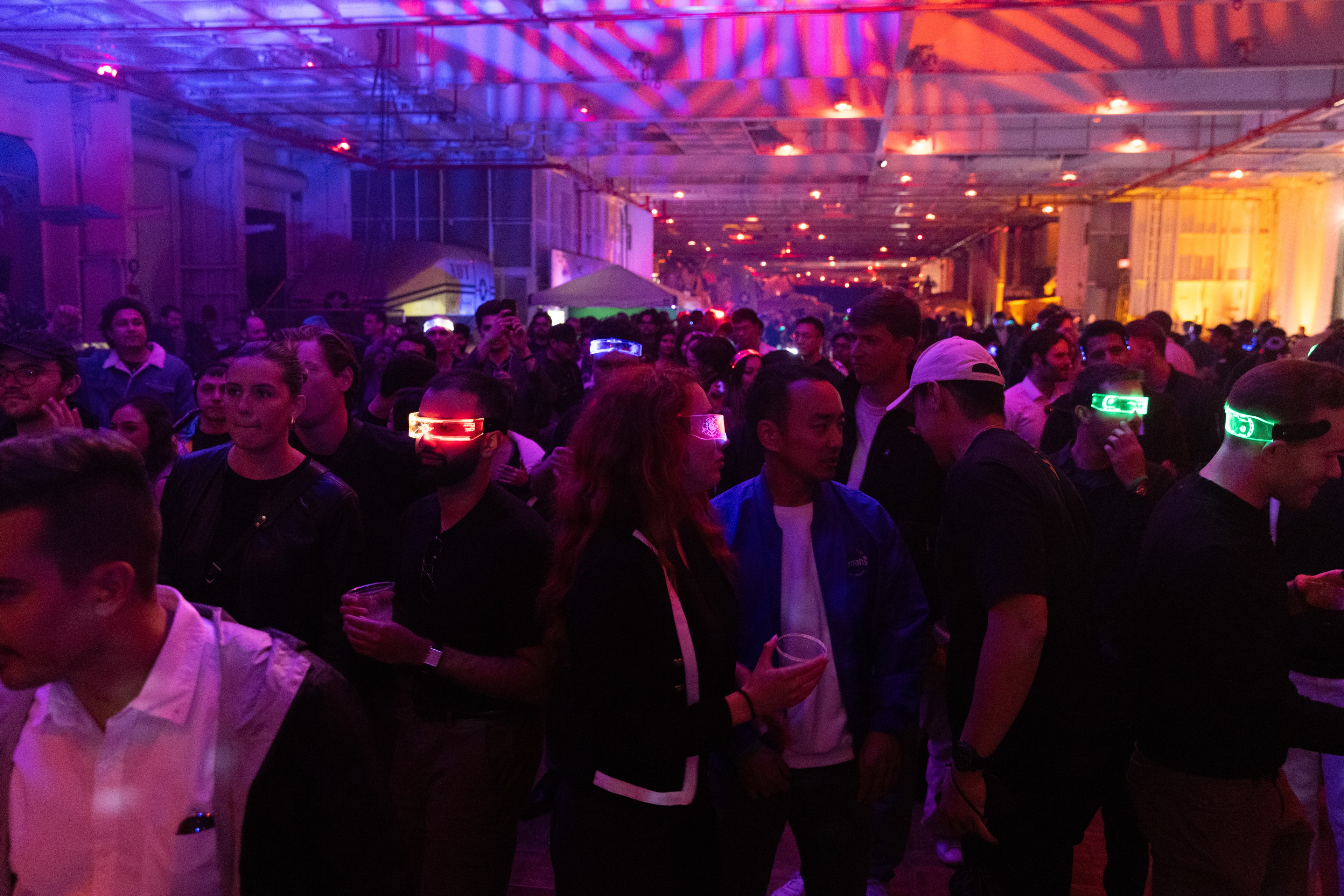 The image shows a crowded indoor event with people wearing colorful LED glasses amid vibrant purple and orange lighting. The setting has an energetic and festive atmosphere.
