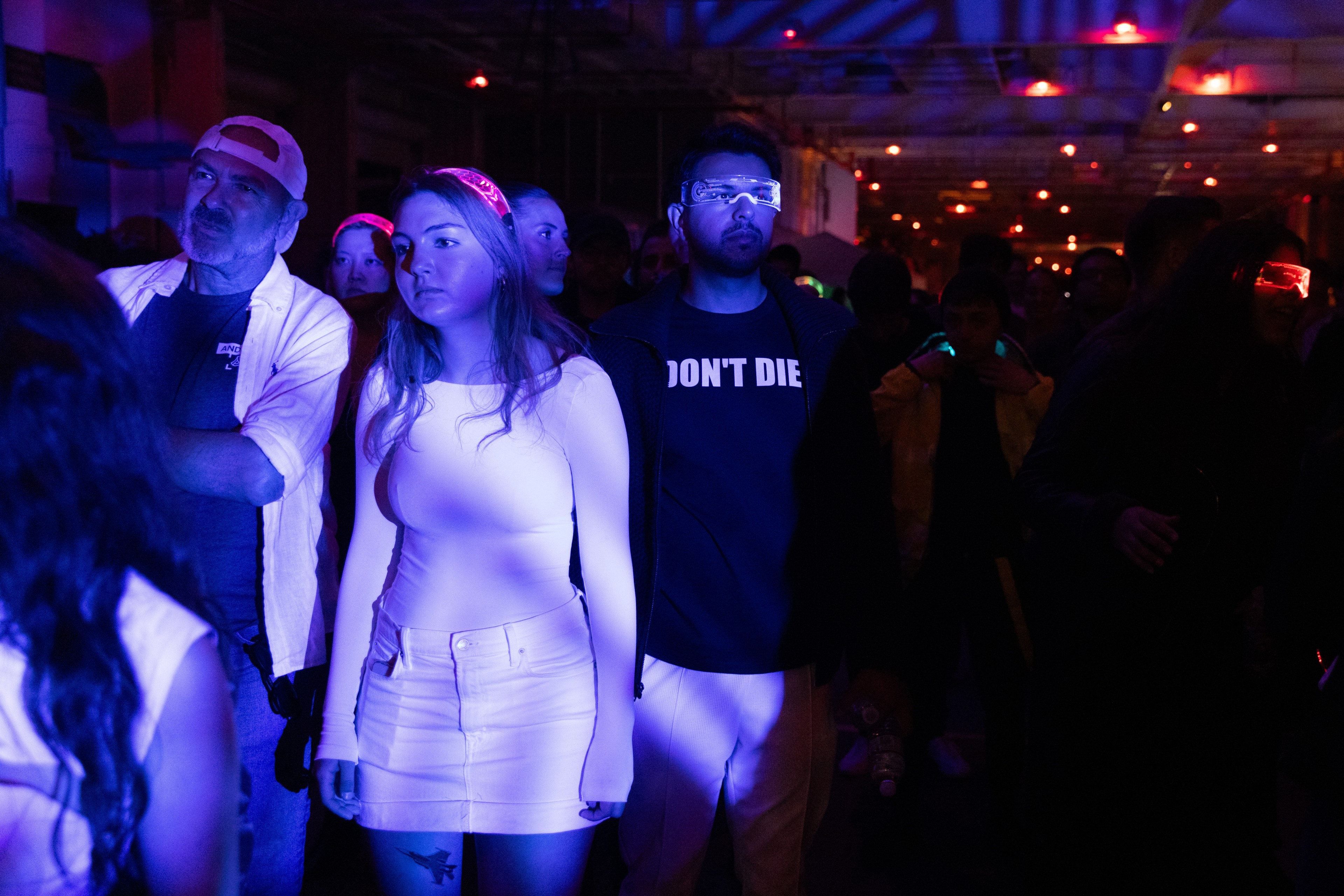 A group of people stand under blue and pink lights at an indoor event, with some wearing light-up accessories and a man in the center wearing a &quot;DON'T DIE&quot; shirt.