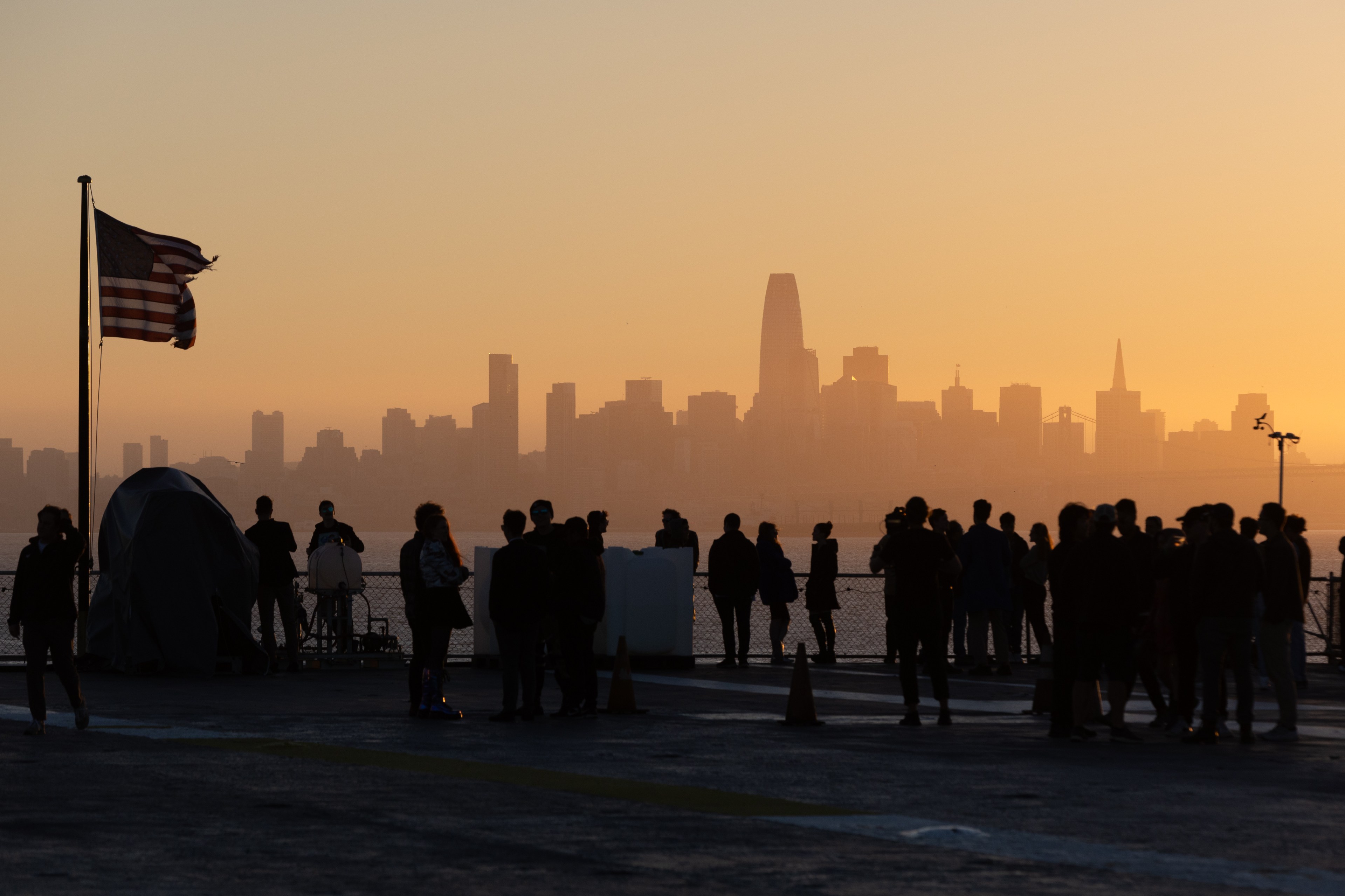 A group of people stand on a dock at sunset, silhouetted against a hazy city skyline. An American flag waves in the breeze on the left side of the image.