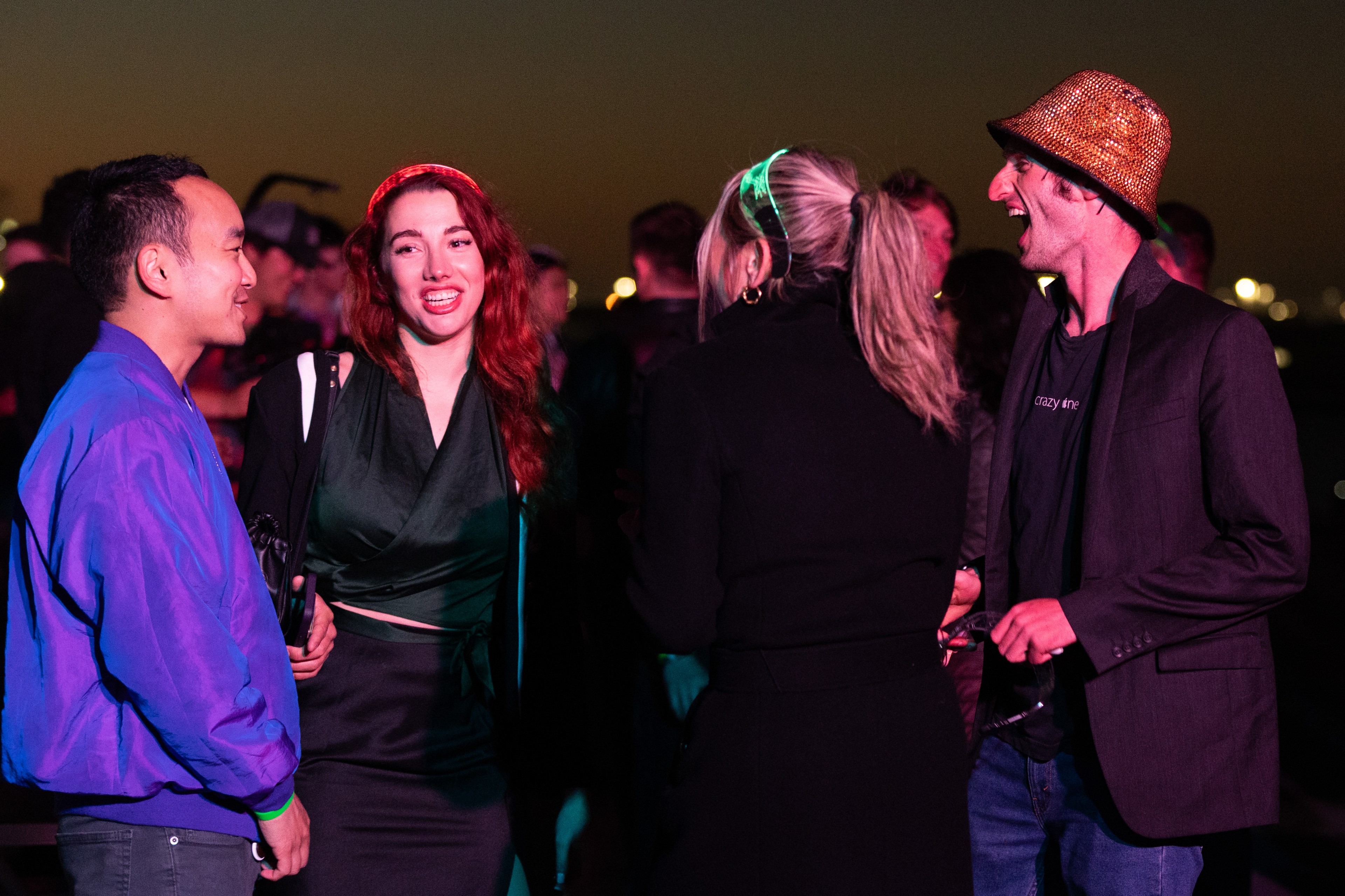 Four people are enjoying a night out, chatting and laughing under dim lights. One woman has red hair, another has a ponytail, a man wears a shiny hat, and another wears a bright jacket.