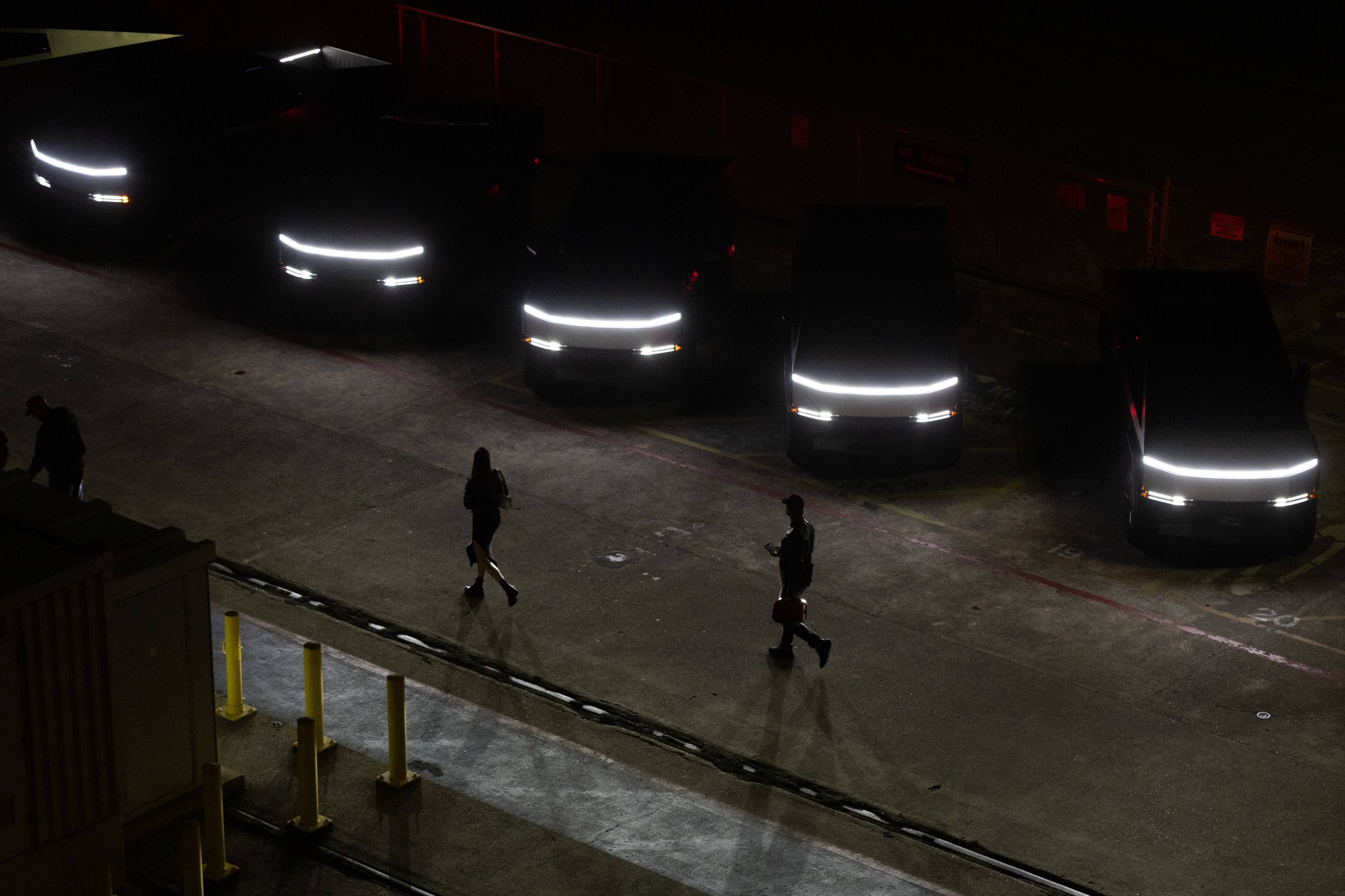 In a dimly lit parking lot, a line of parked vehicles with bright horizontal headlights faces forward while two people walk in the foreground, casting long shadows.