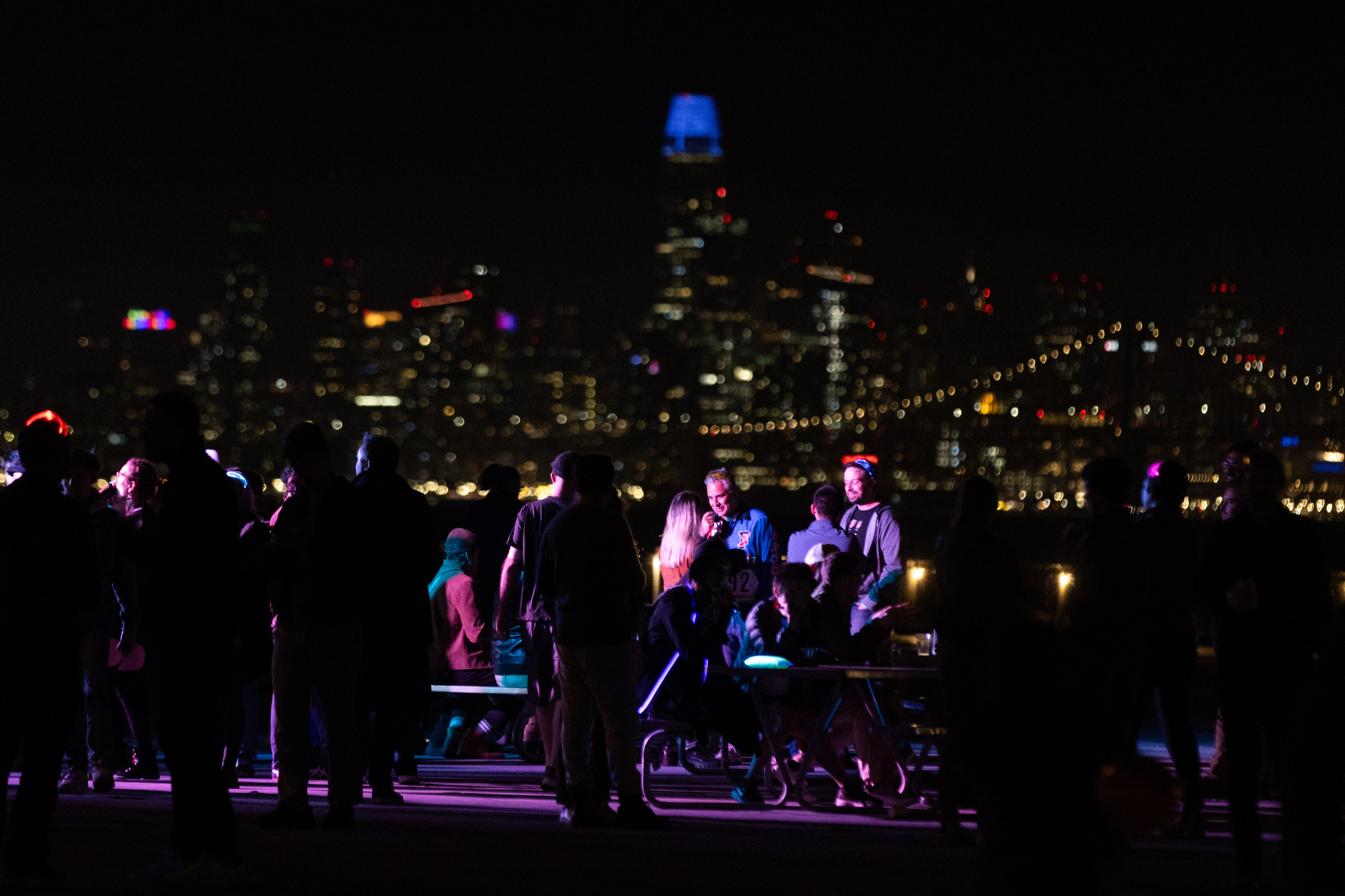 A group of people are socializing at night, partially illuminated by colored lights. The background features a vibrant, glowing city skyline with a visible bridge.