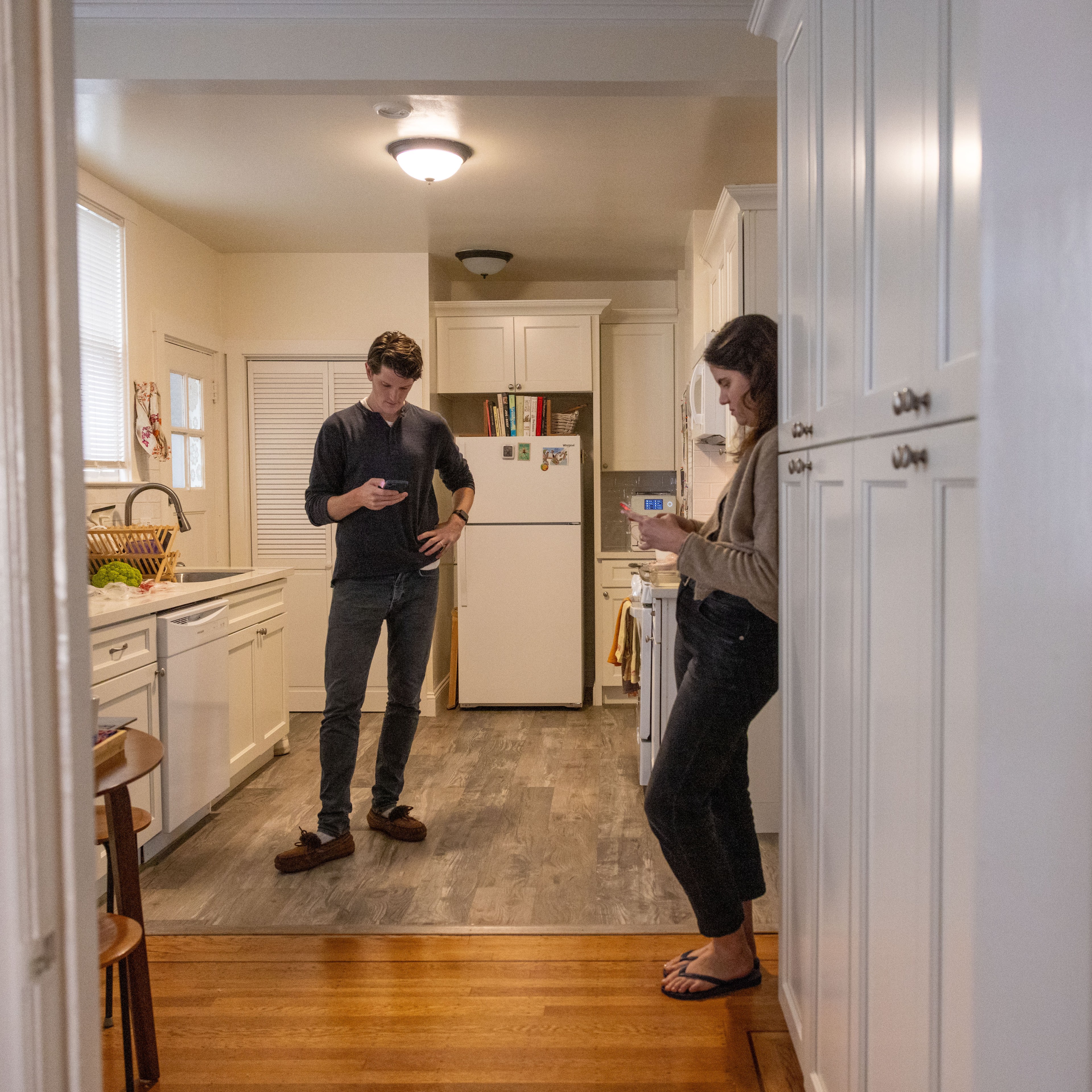 Two people are standing and looking at their phones in a clean kitchen; one leans against a wall, the other stands with one hand on their hip.