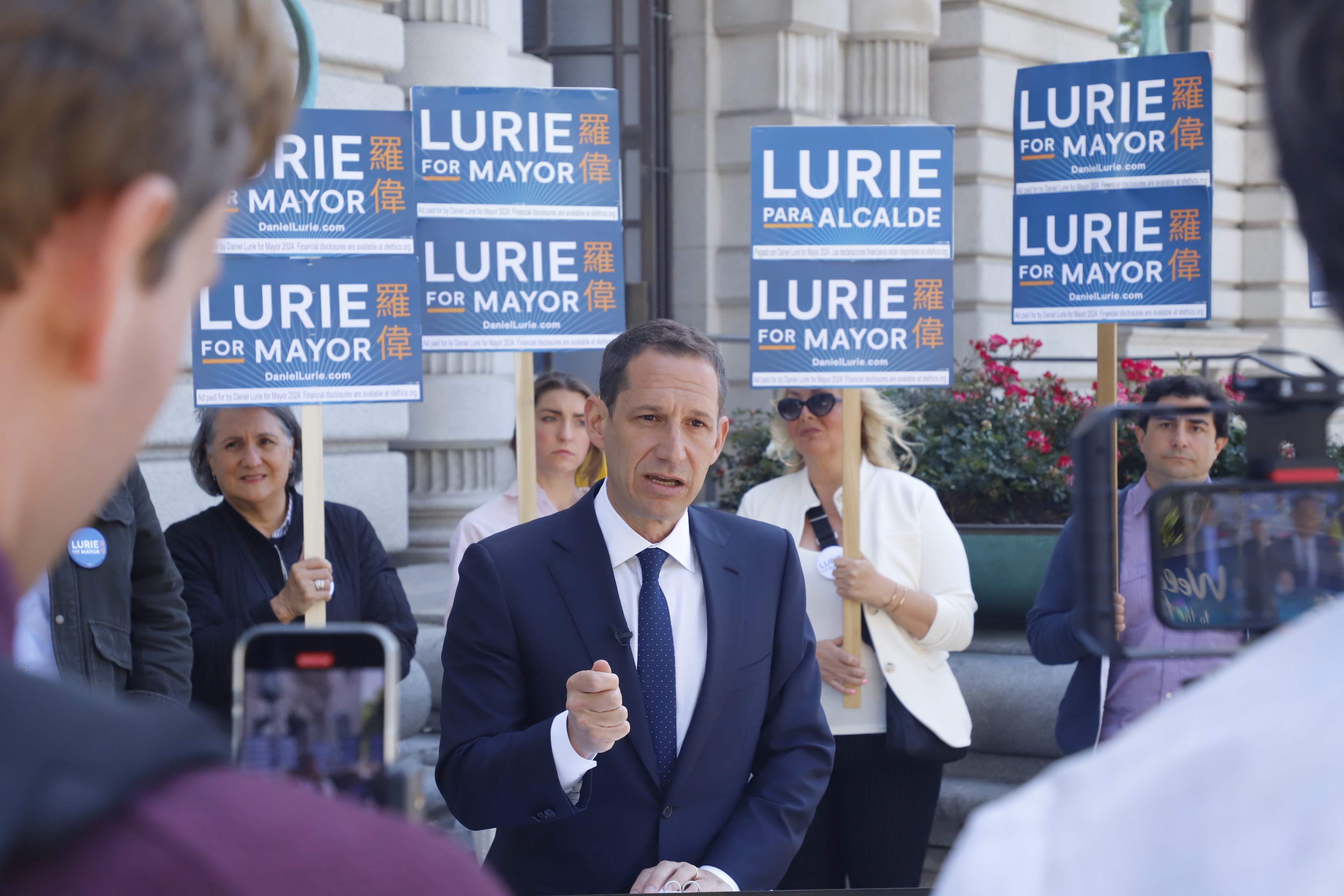 A man in a suit speaks at a podium, surrounded by people holding &quot;Lurie for Mayor&quot; signs. The group stands outside a building, with a few recording the event.