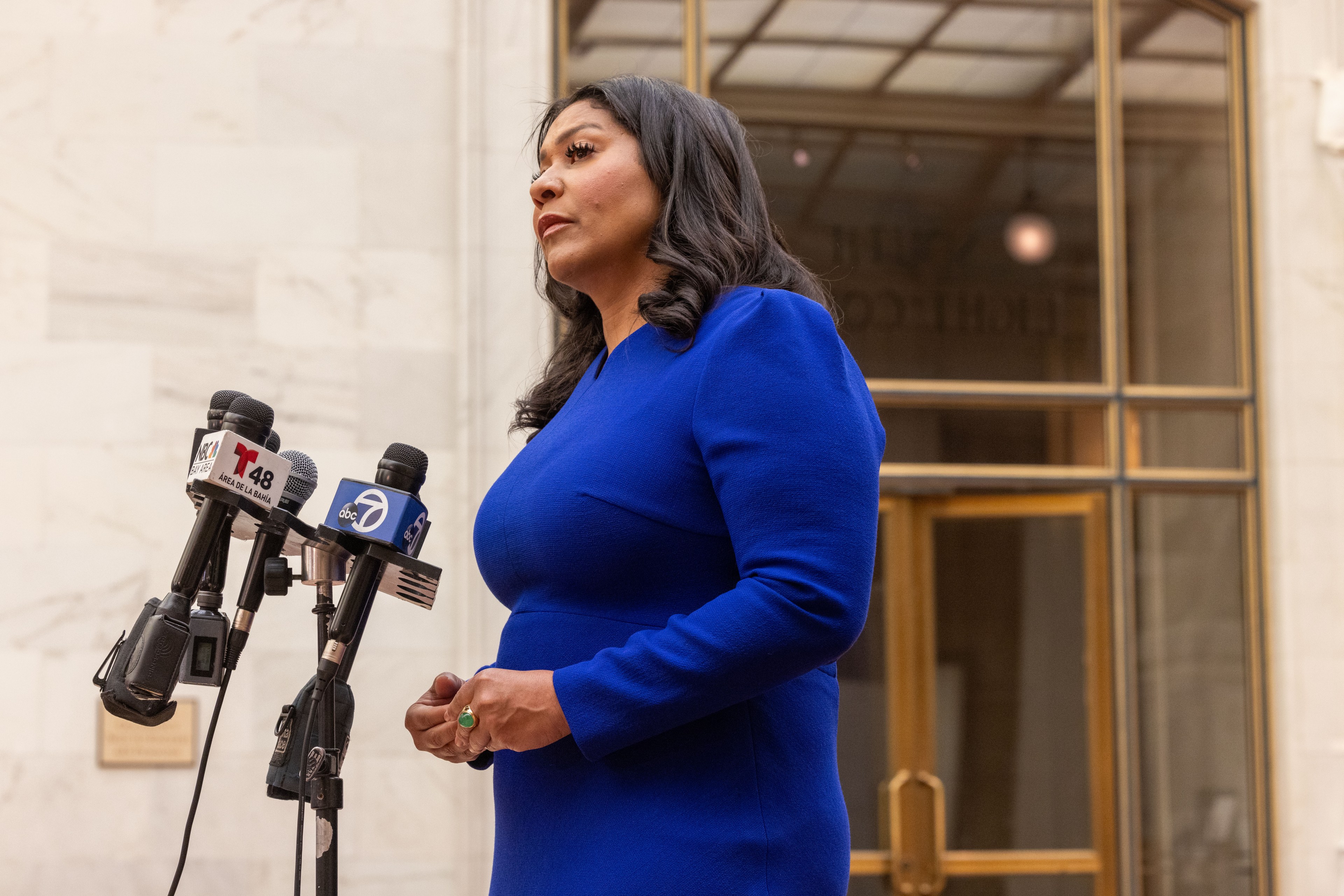 A woman in a blue dress stands before microphones at a press conference at City Hall.