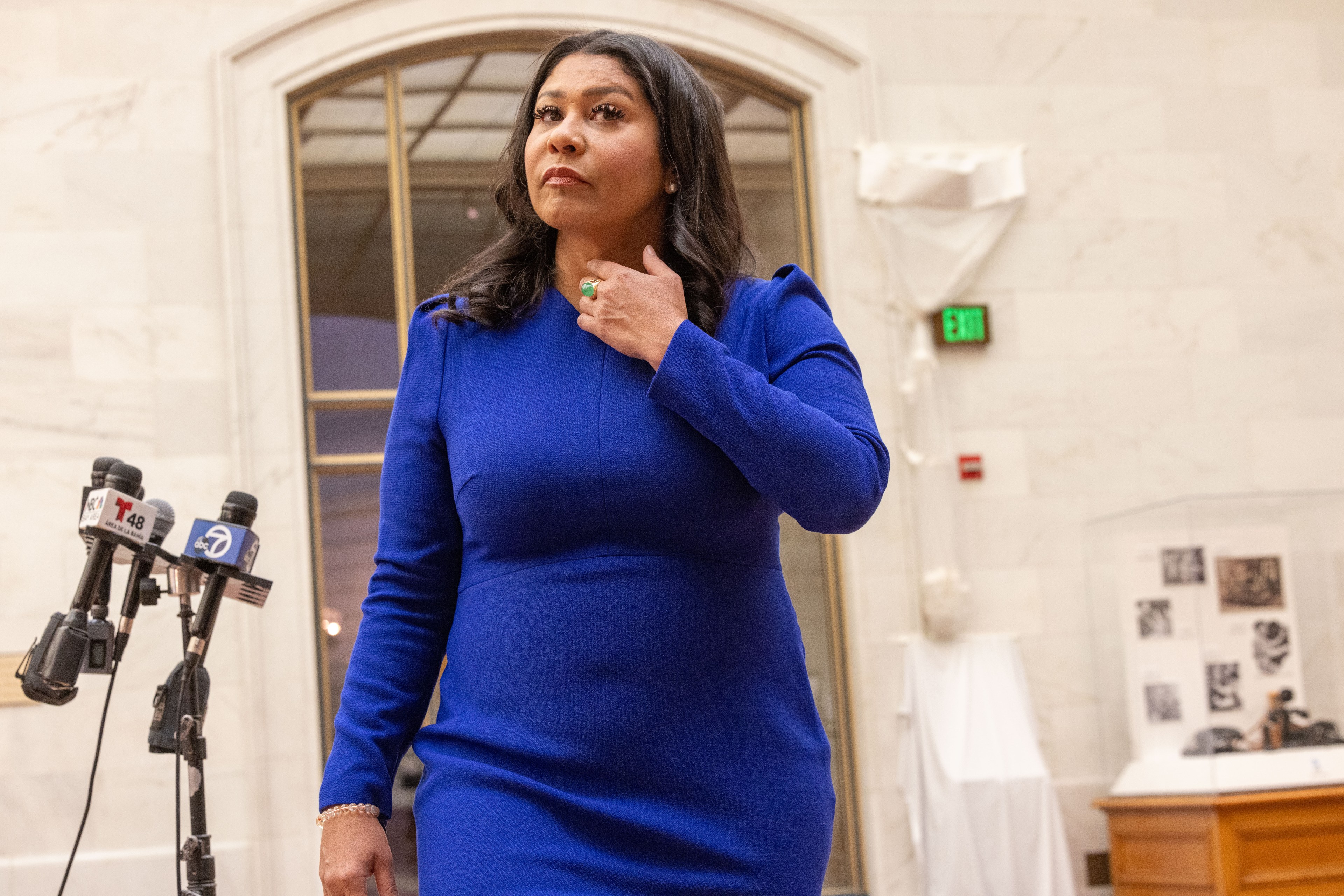 Mayor London Breed in a blue dress stands near microphones, adjusting her necklace. The background features a marble wall, an arched window, and display cases.