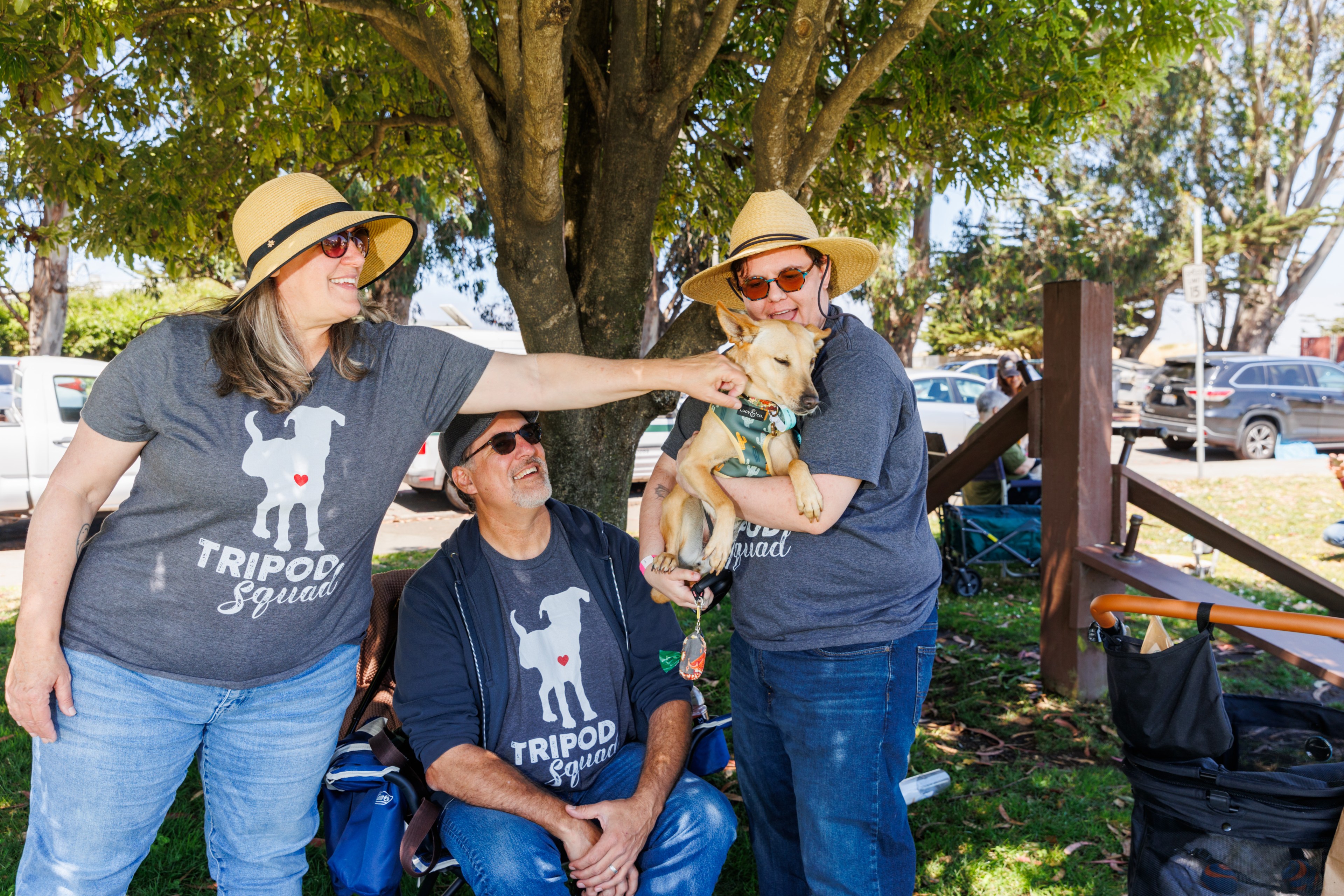 Three people wearing &quot;Tripod Squad&quot; shirts and hats are gathered under a tree. One woman holds a light-colored dog, and another woman reaches out to pet it.