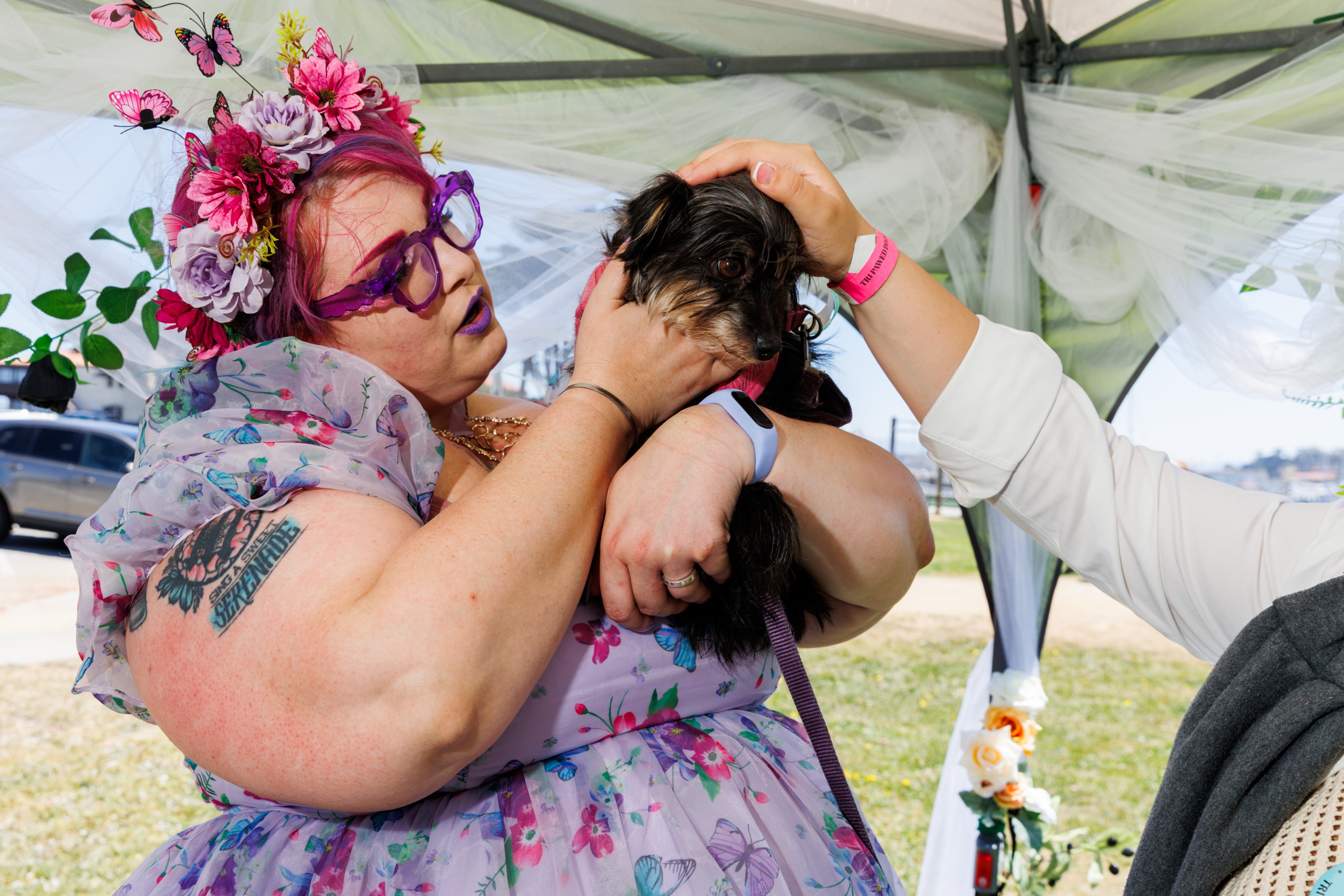A person with pink hair, purple glasses, and a floral headdress holds a small dog while another person pets the dog under a decorated canopy outdoors.