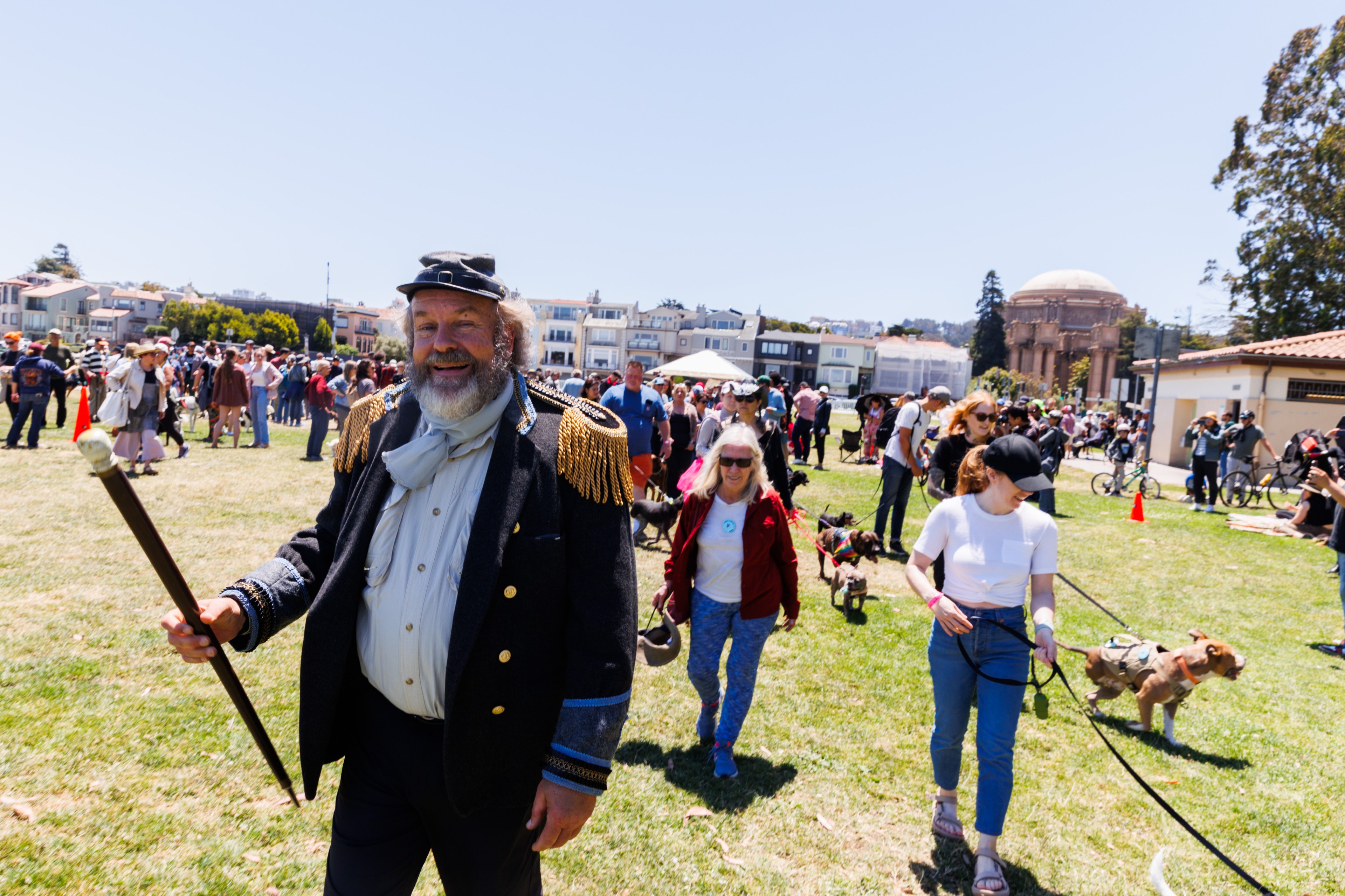 A bearded man in 19th-century attire holding a staff leads a crowd in a park. People walk dogs, and buildings are visible in the background under a clear sky.