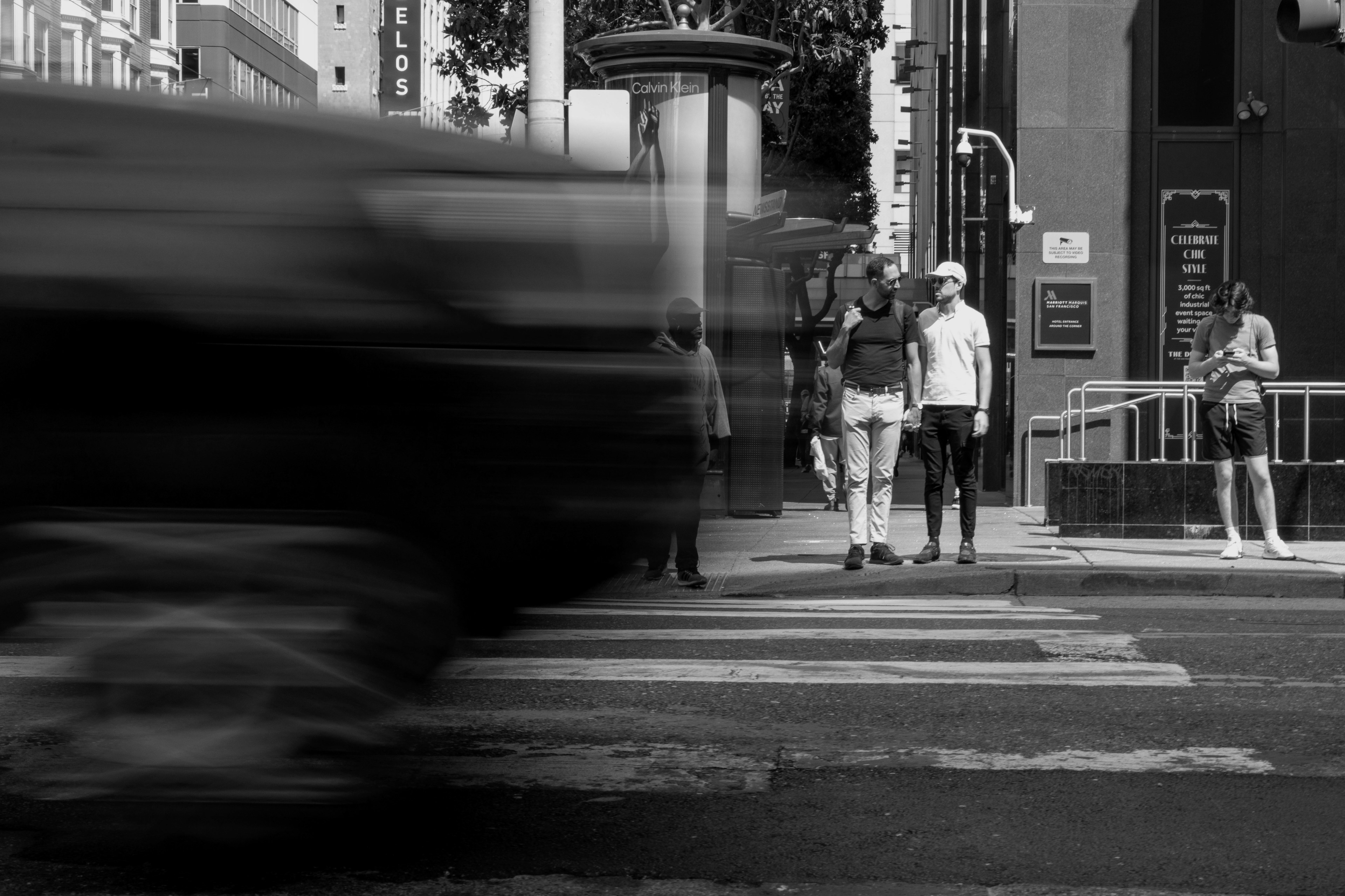 In a city street scene, several people wait to cross while a blurred vehicle speeds past in the foreground, creating a dynamic contrast between motion and stillness.