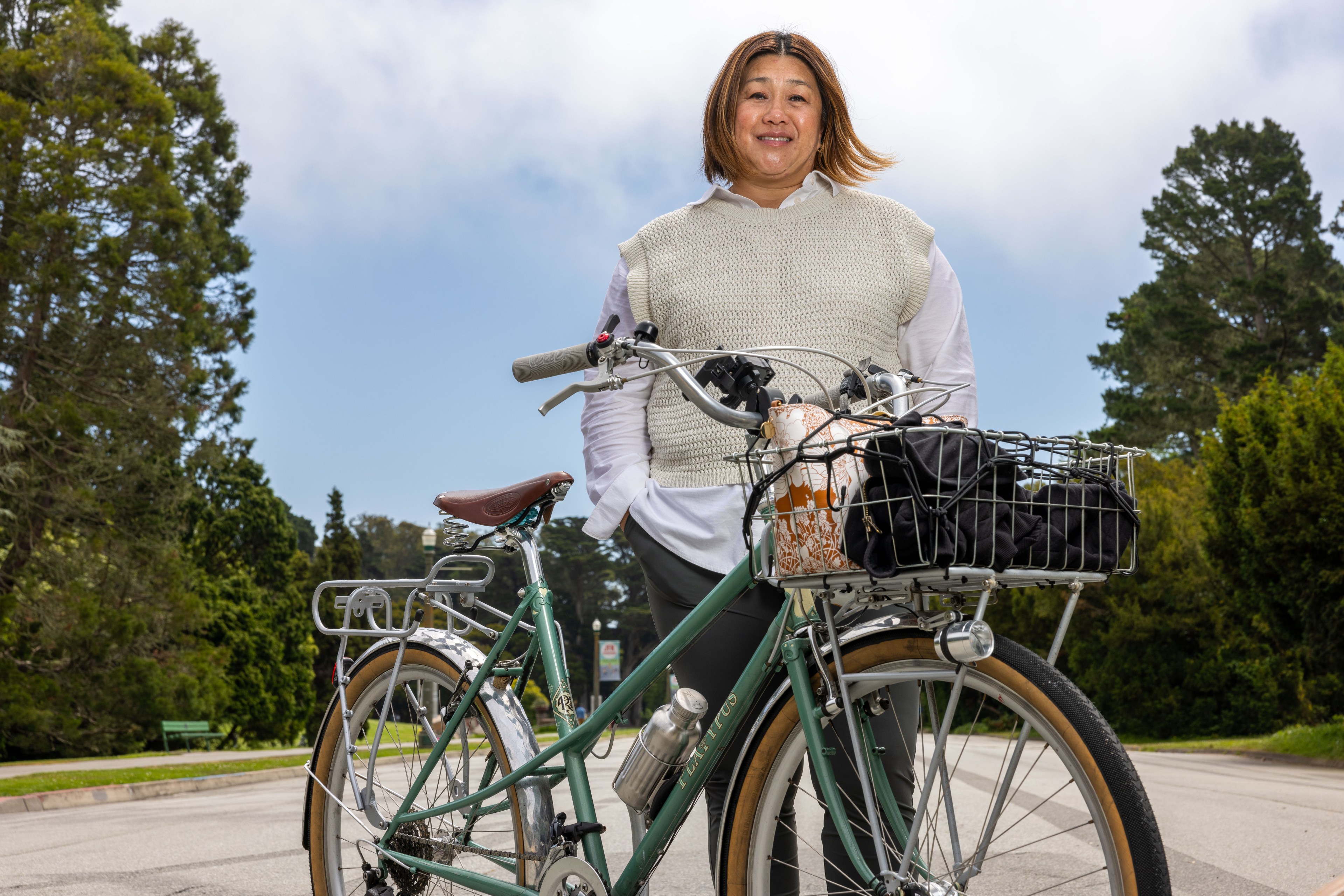 A person stands smiling beside a green bicycle with a loaded front basket on a tree-lined street, wearing a white shirt and light-colored sweater vest.
