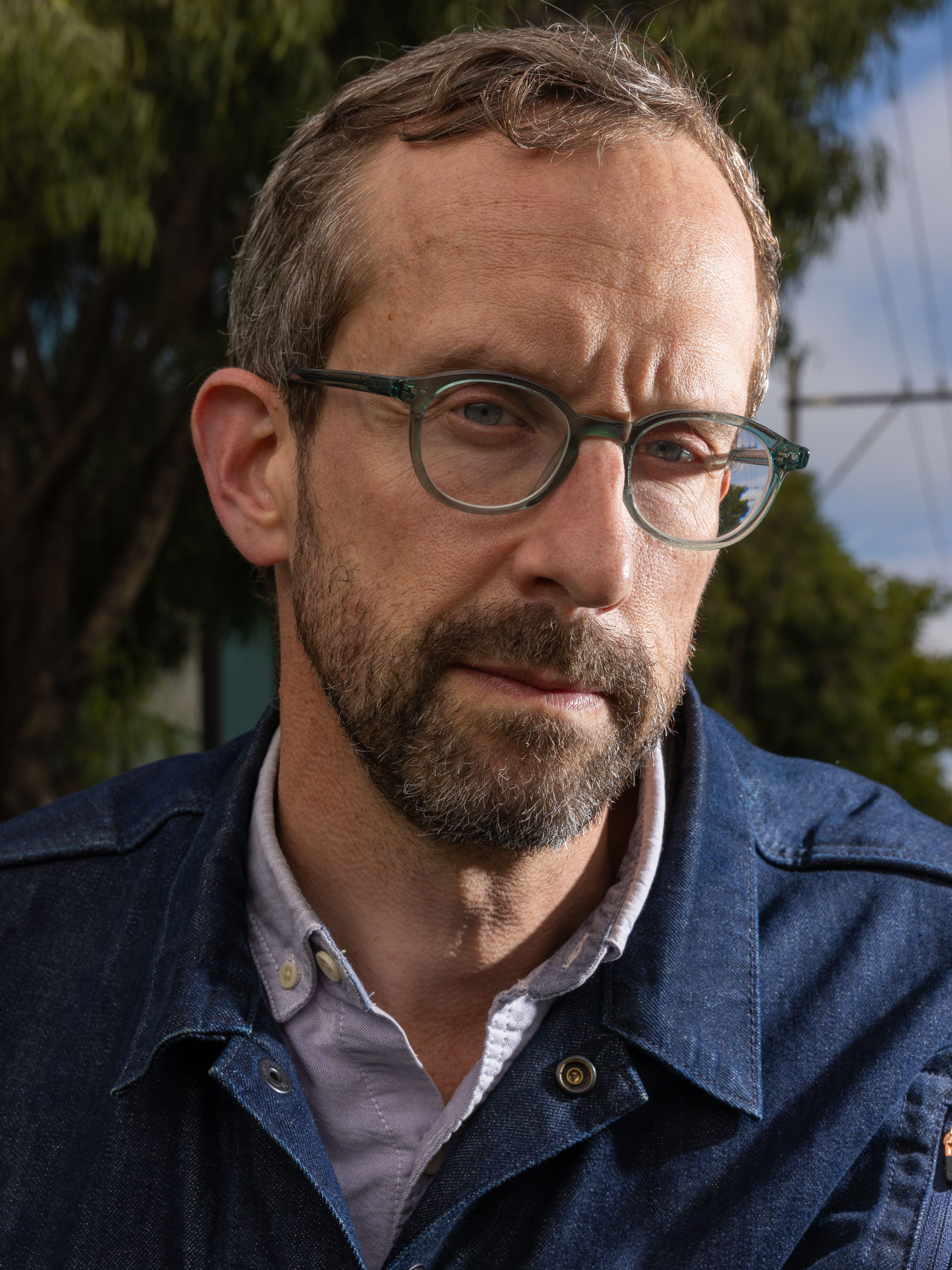 A man with glasses and a beard, wearing a denim jacket over a light-colored shirt, stands outdoors. Trees, a building, and power lines are visible in the background.
