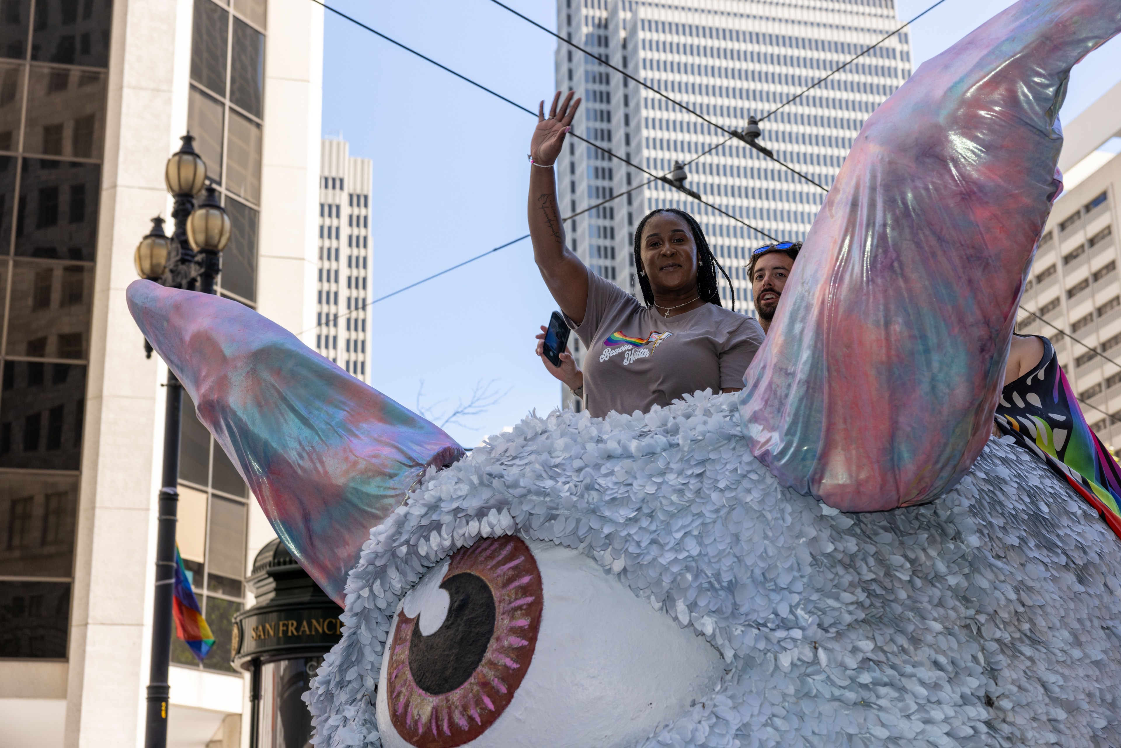 Two people wave from atop a large, colorful creature float with big ears and an eye, set against a cityscape of tall buildings and street cables.