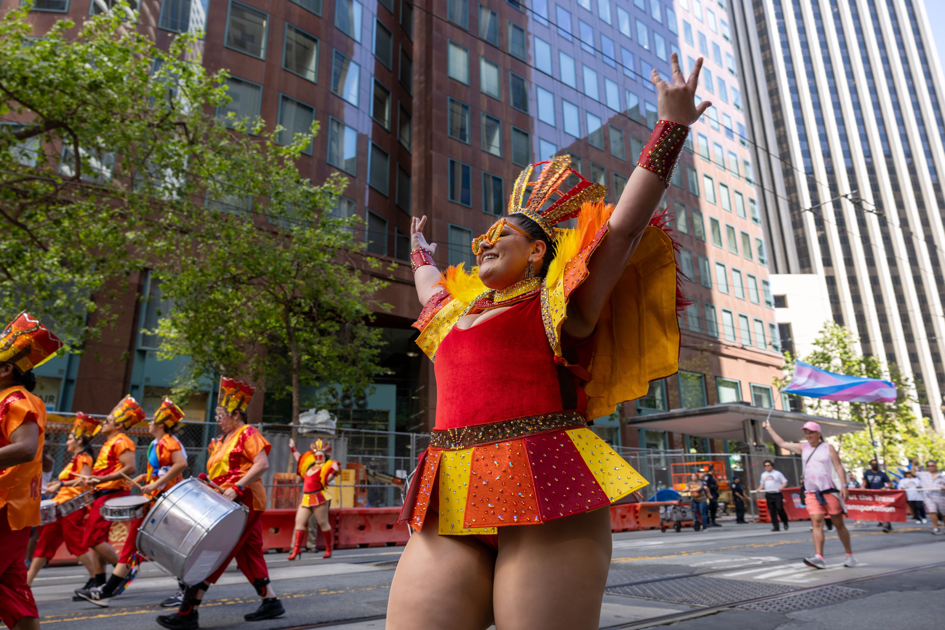 A person in a vivid red and yellow costume with arms raised is leading a parade. Drum players march behind, and tall city buildings and trees form the background.
