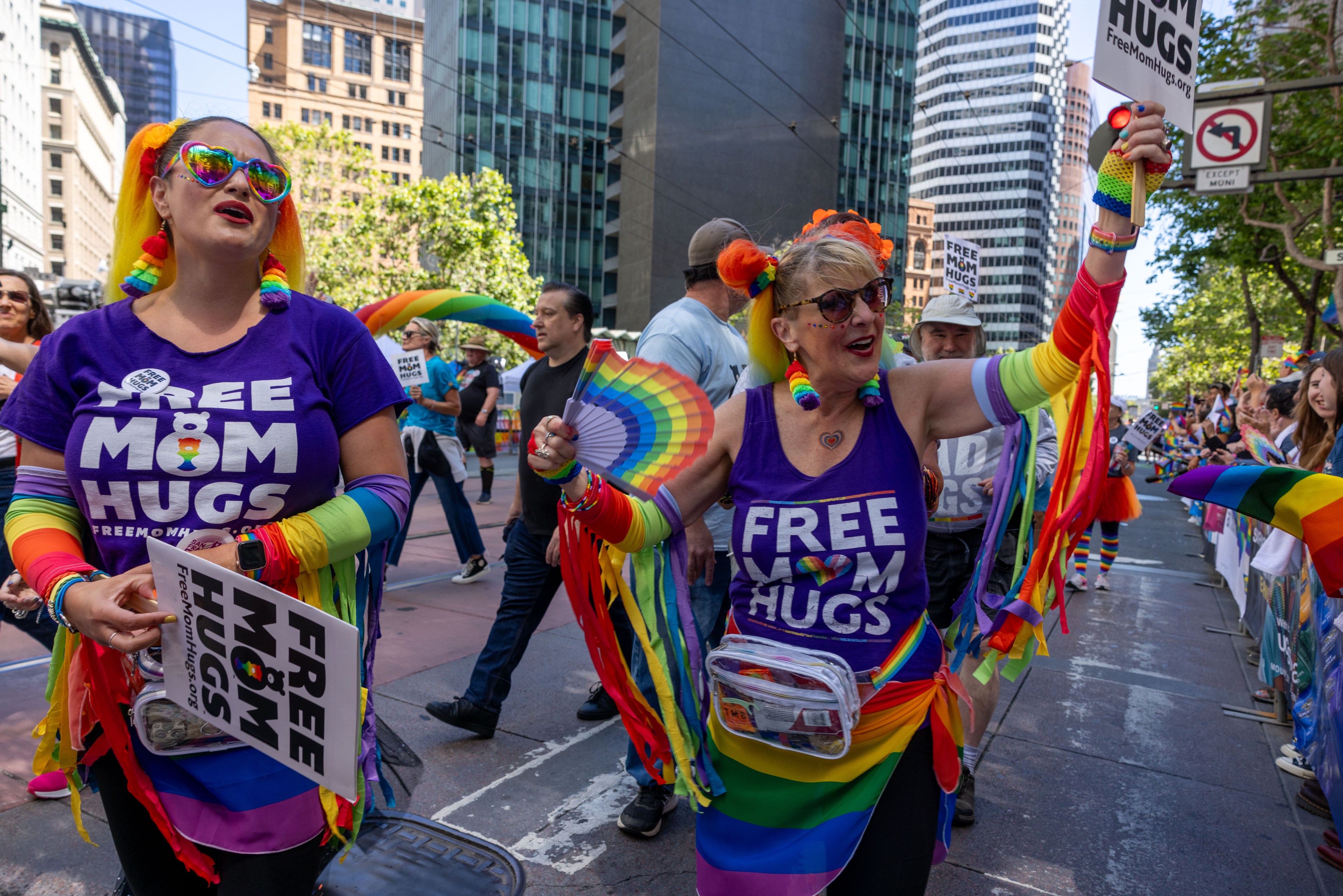 Two women in colorful outfits and &quot;Free Mom Hugs&quot; shirts are joyfully participating in a parade, holding rainbow fans and signs, surrounded by buildings and a cheering crowd.