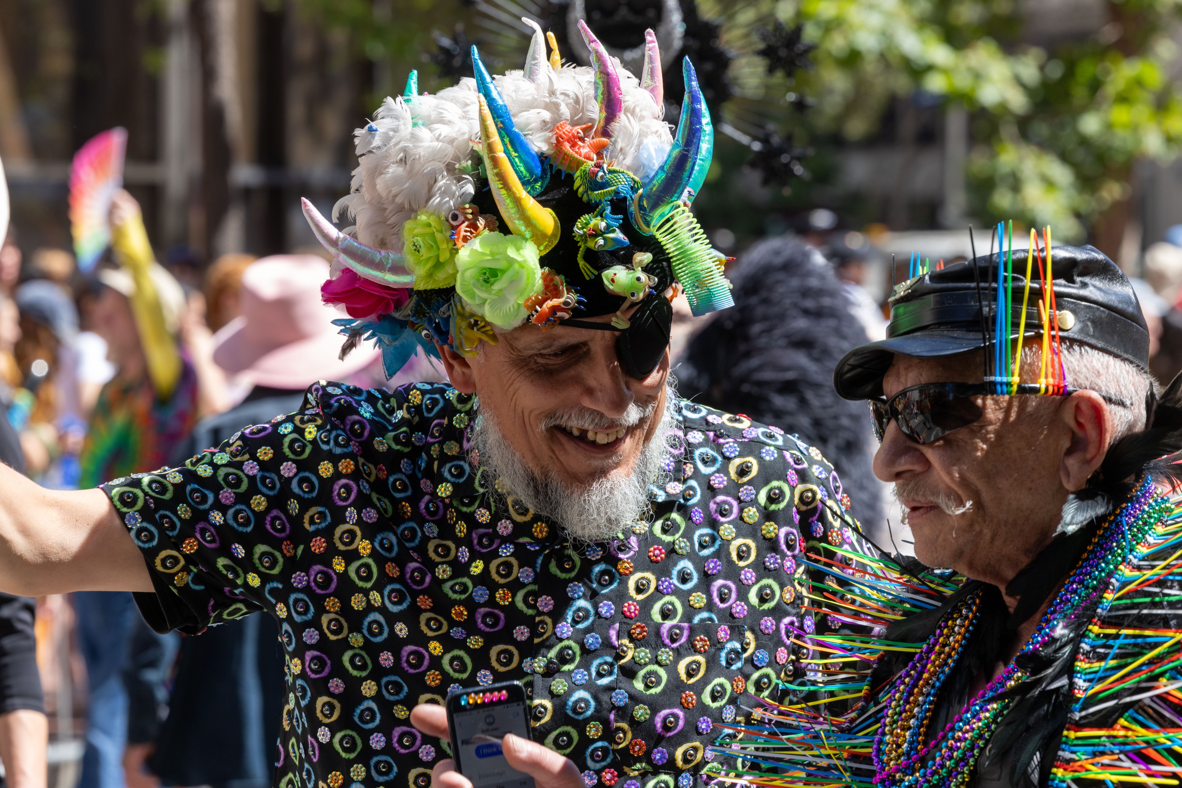 Two men in colorful, eccentric outfits are smiling and talking. One has a hat with multicolored horns and flowers; the other wears a hat with rainbow decorations.