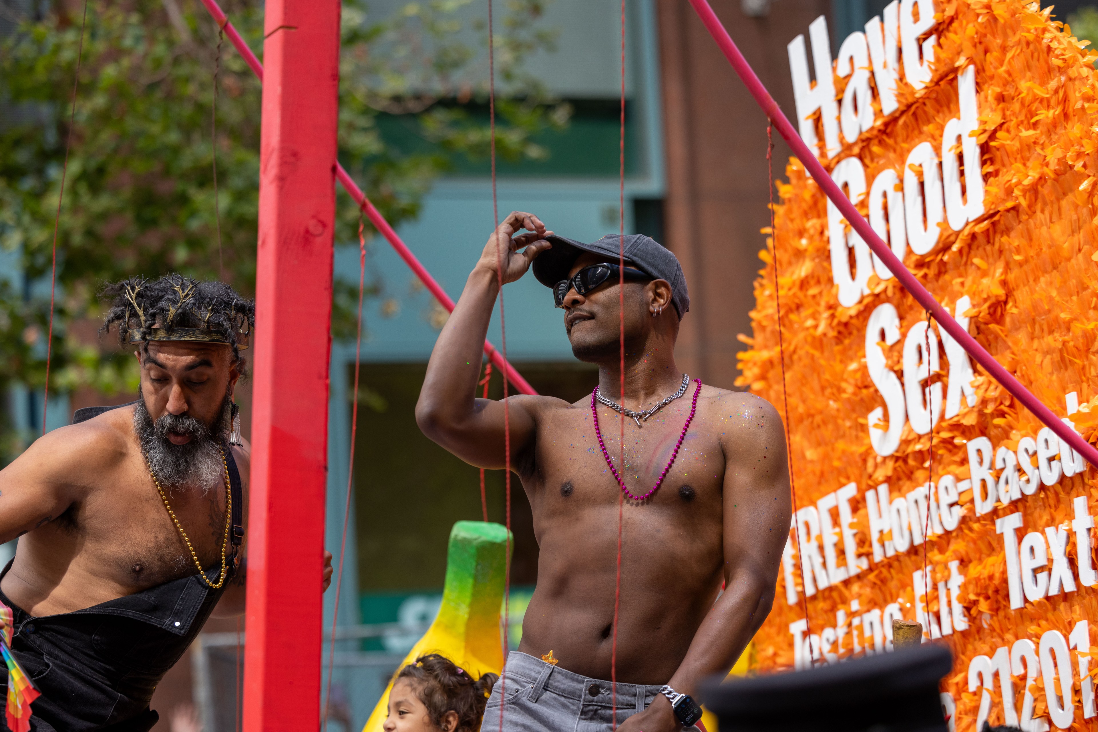 Two shirtless men are celebrating on a float. One wears a flower crown and the other a gray cap and beads. The float has an orange sign with bold white text.