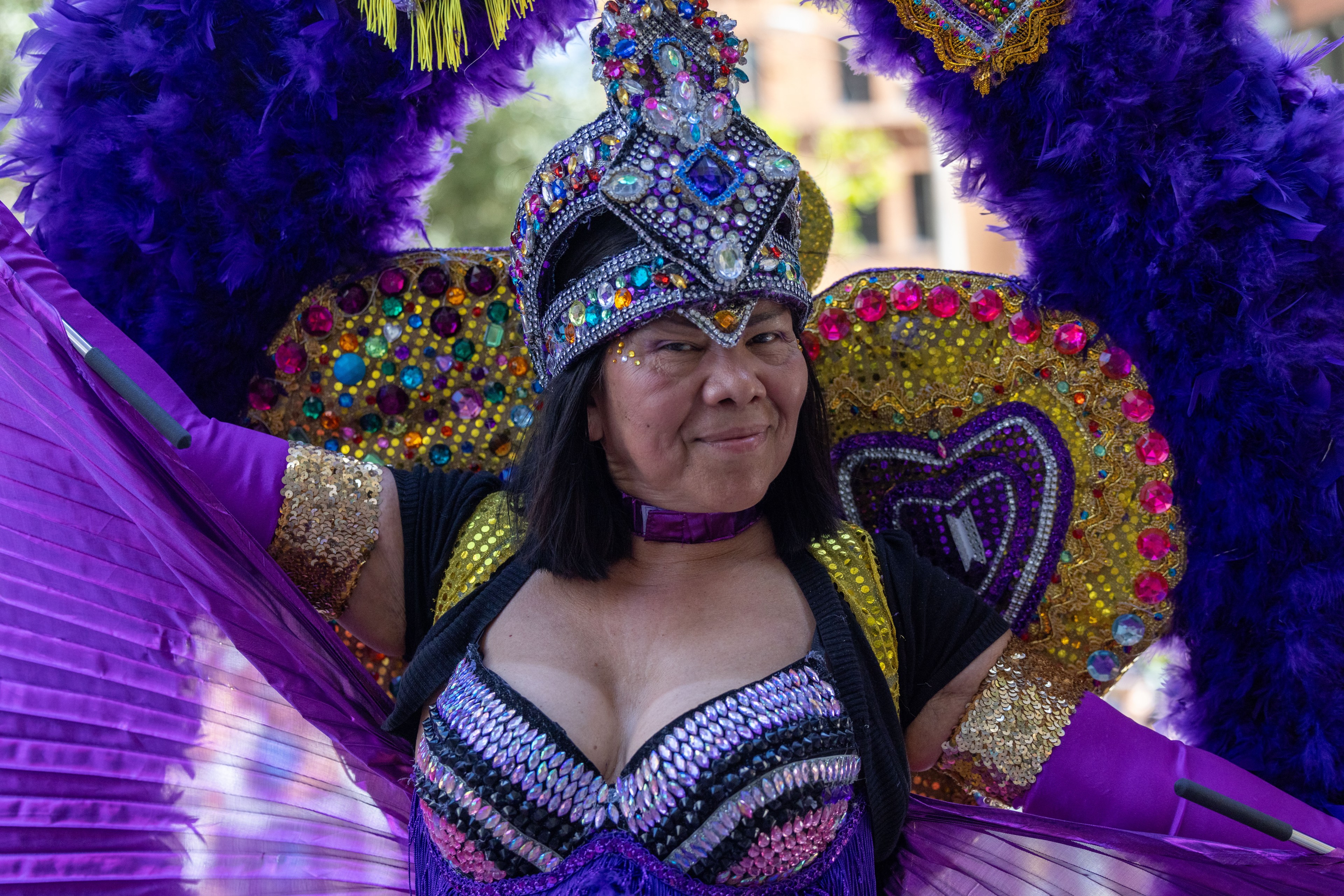 A person is wearing an embellished, colorful costume with purple feathers, sequins, and jewels, smiling confidently. They have a matching elaborate headpiece and winged decorations.