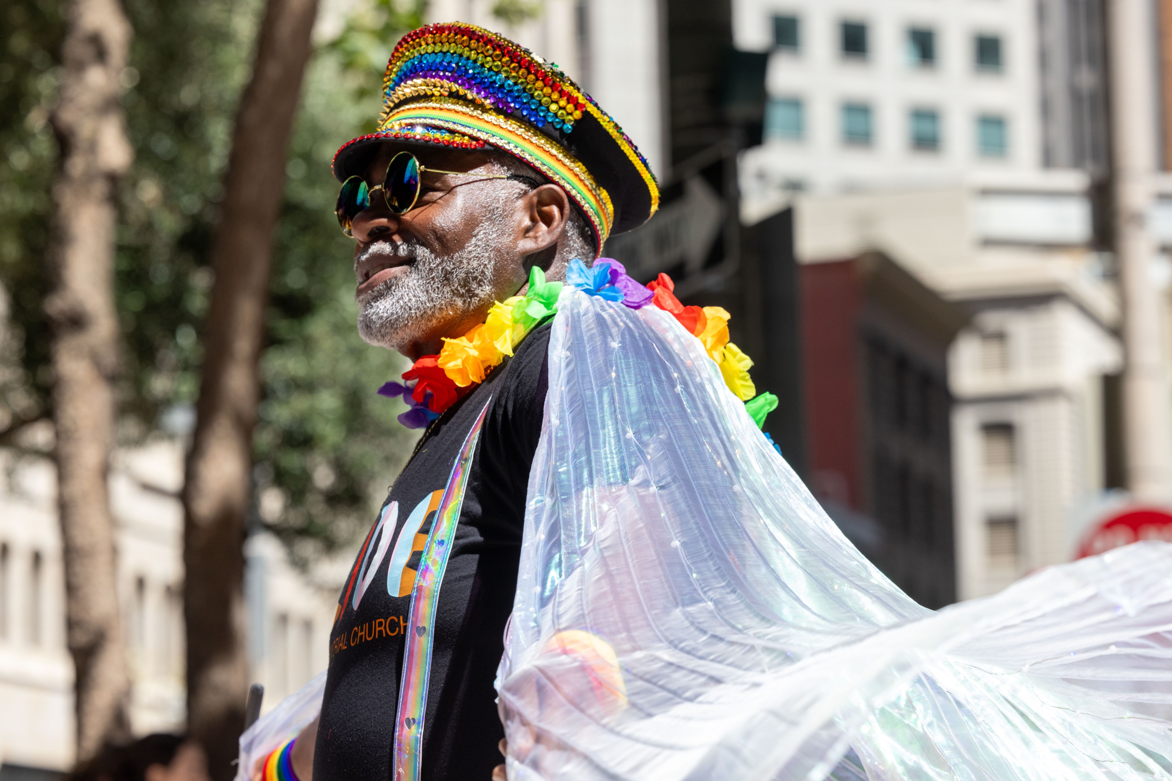 A man wears a colorful, sequin-covered hat and a rainbow lei. He has sunglasses on, a beard, and a black T-shirt with rainbow suspenders, draped in a sheer, iridescent fabric.
