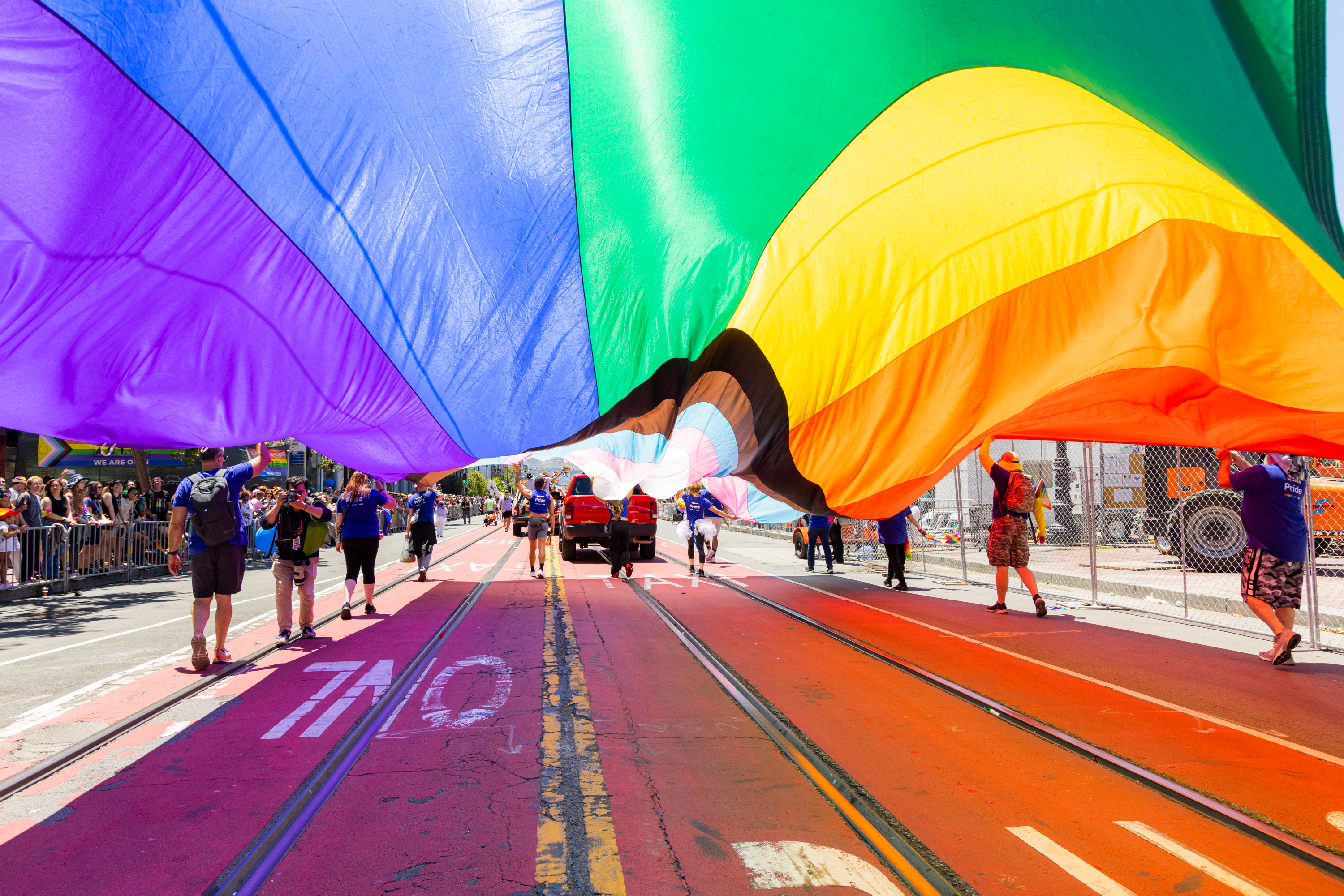 People march under a large rainbow flag during a parade. The vibrant colors cover the street, with spectators watching from the sides.