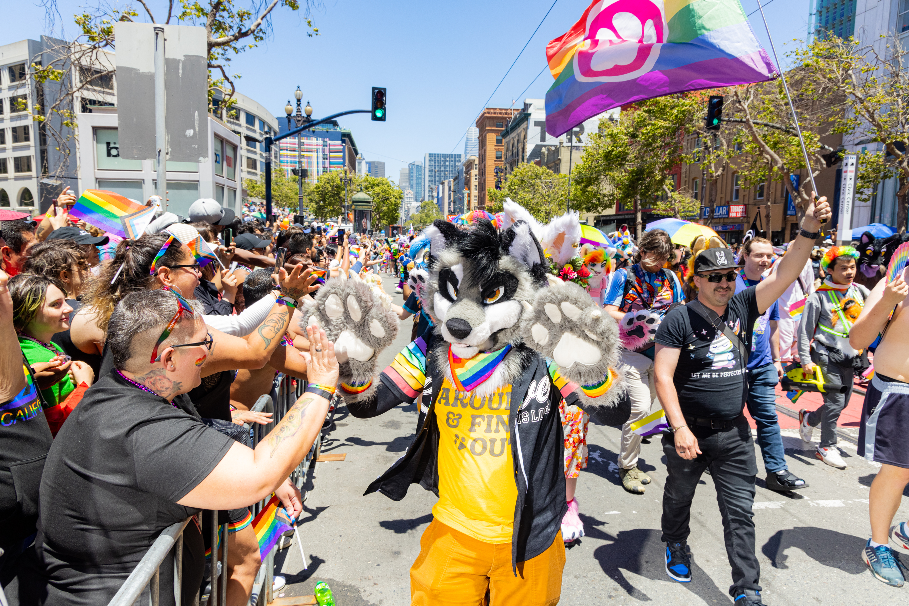 A colorful parade features a person in a wolf costume, surrounded by a joyful crowd waving rainbow flags, high-fiving attendees in a sunny urban setting.