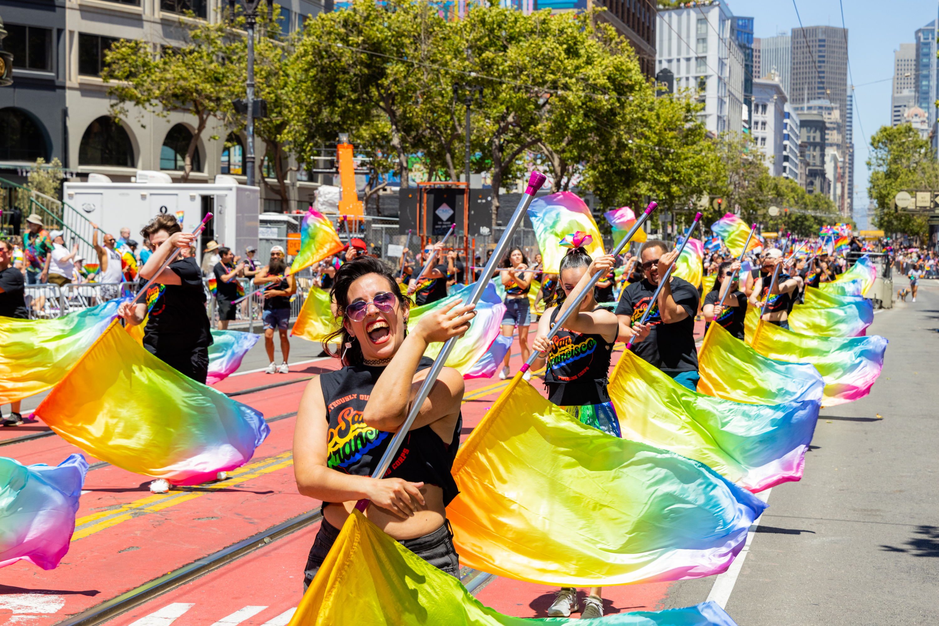 A group of people, joyfully waving colorful rainbow flags, parades down a city street lined with buildings and spectators under a bright, sunny sky.